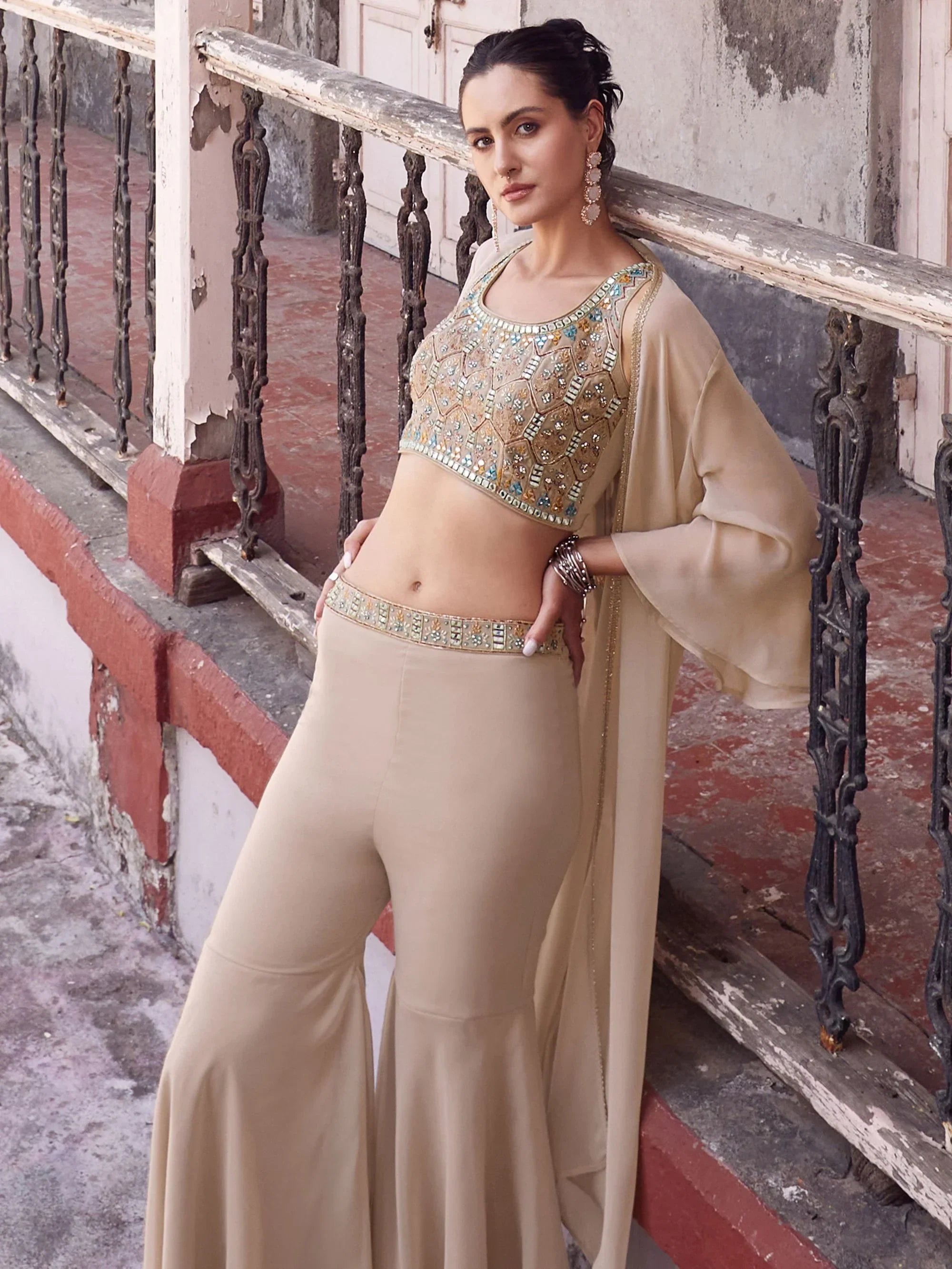 Woman in beige embellished crop top and flared pants, wearing ethnic jewelry, leaning on railing