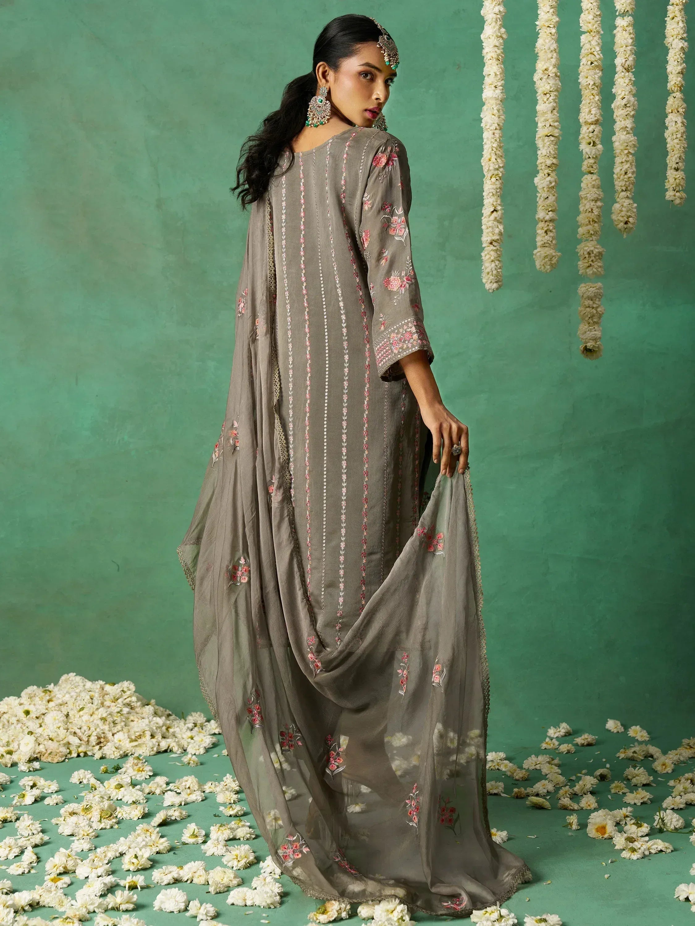 Grey floral embroidered salwar kameez with dupatta, styled with traditional jewelry against a green floral backdrop.
