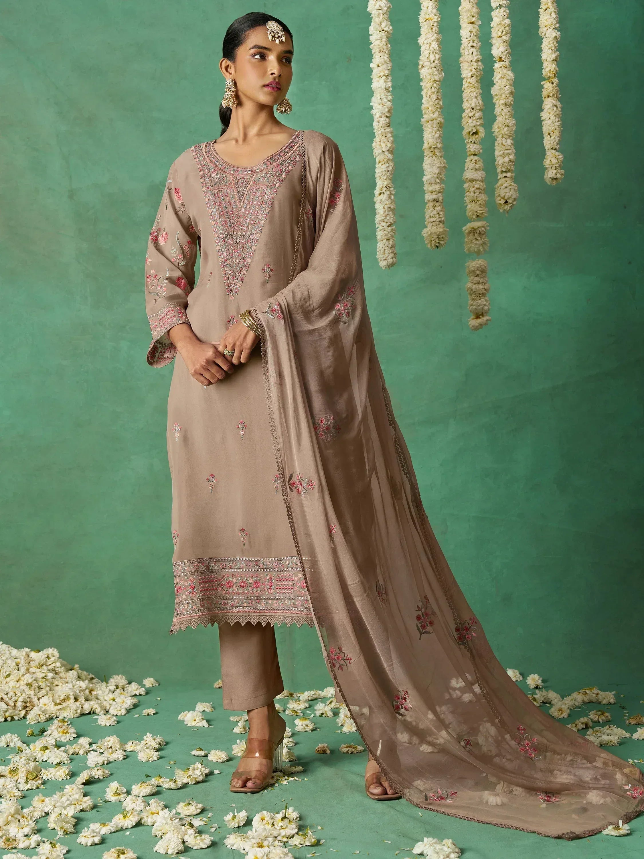 Beige embroidered salwar kameez with floral motifs, dupatta, and traditional jewelry against green floral backdrop