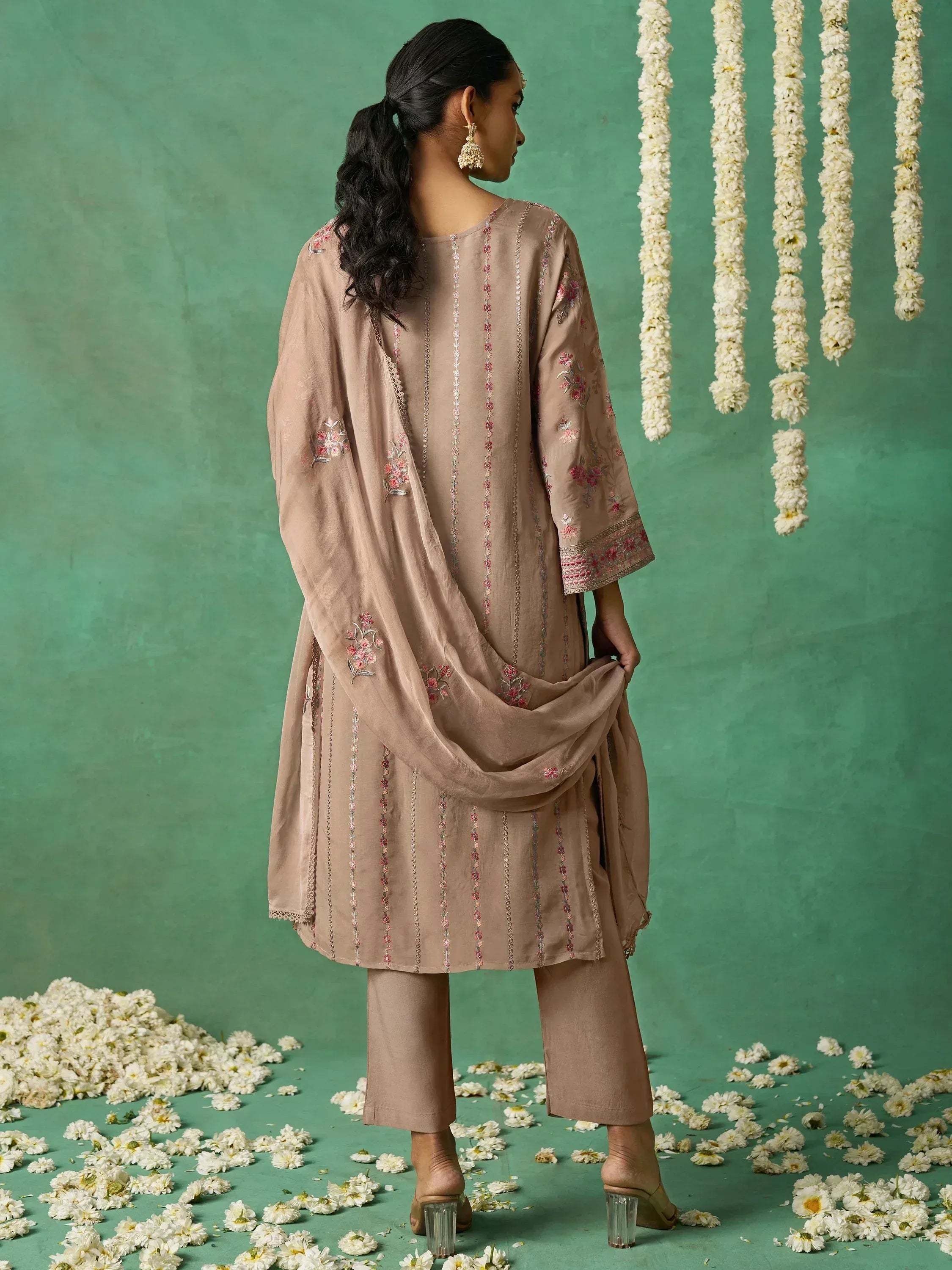 Beige embroidered salwar kameez with floral details, styled with dupatta against green backdrop with white flowers