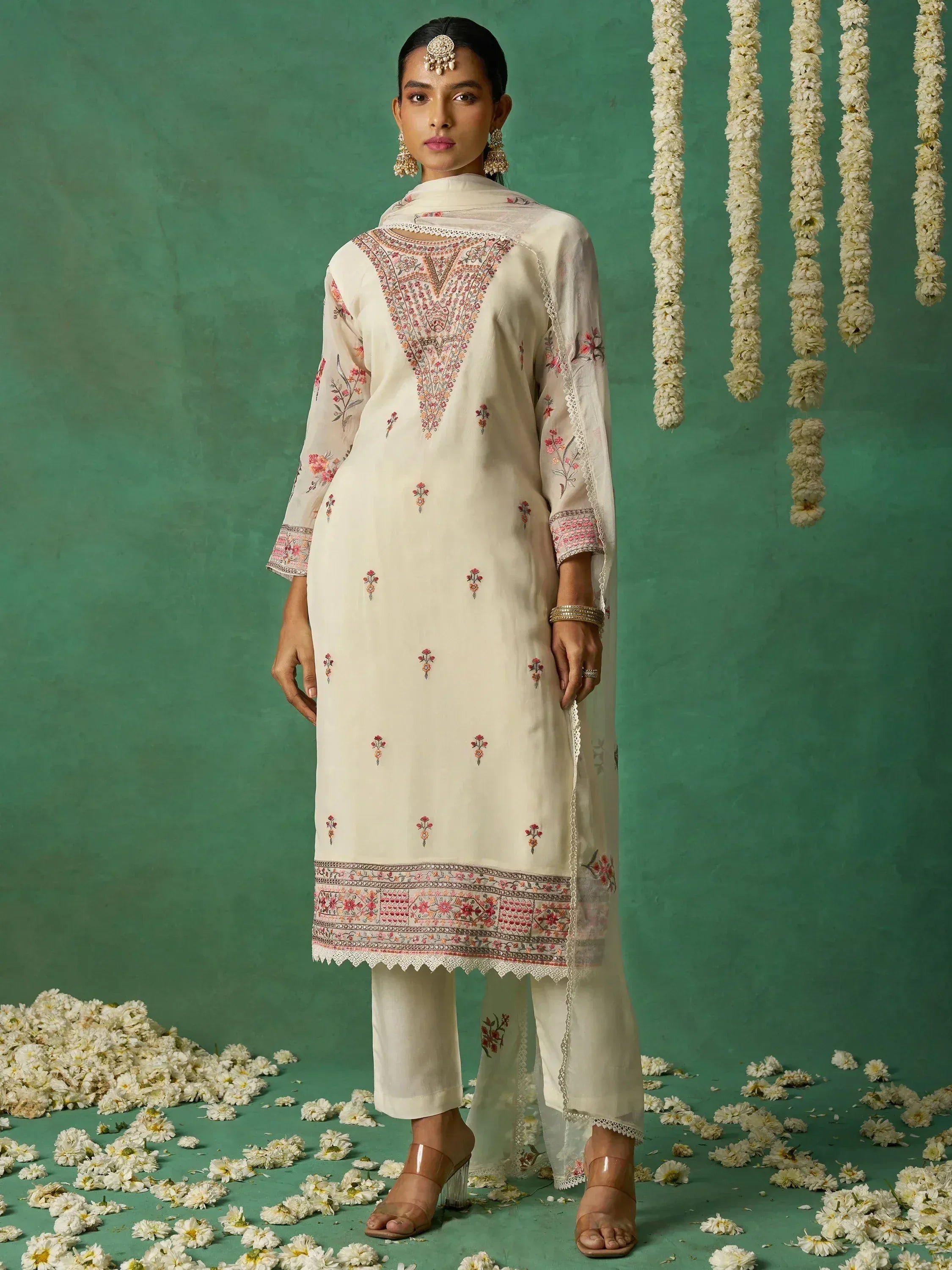 Cream embroidered salwar kameez with floral motifs, dupatta, and traditional jewelry against green backdrop with flowers