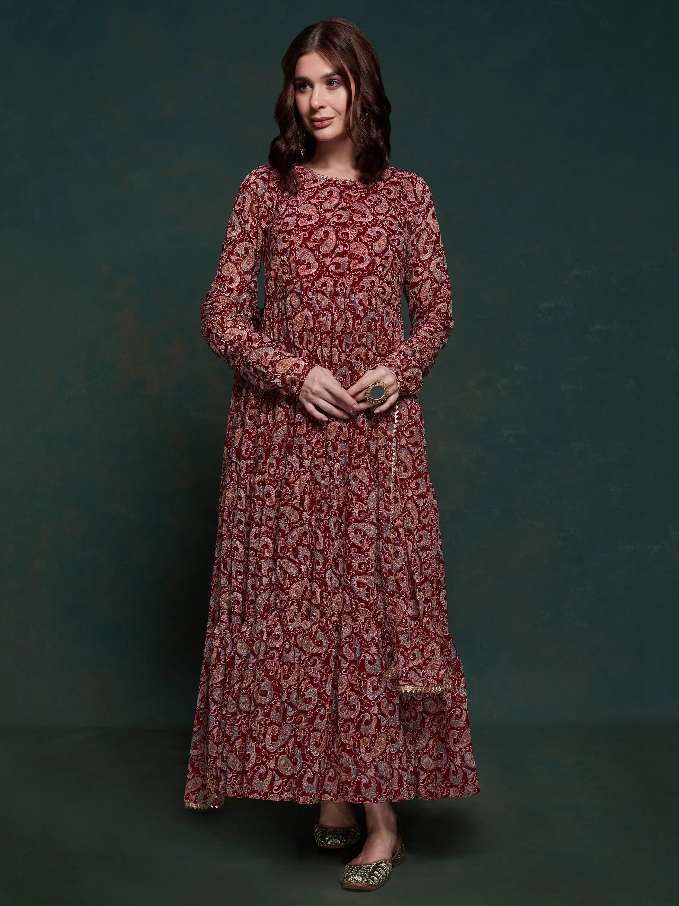 Woman in a maroon paisley print ethnic gown with long sleeves, standing against a dark background