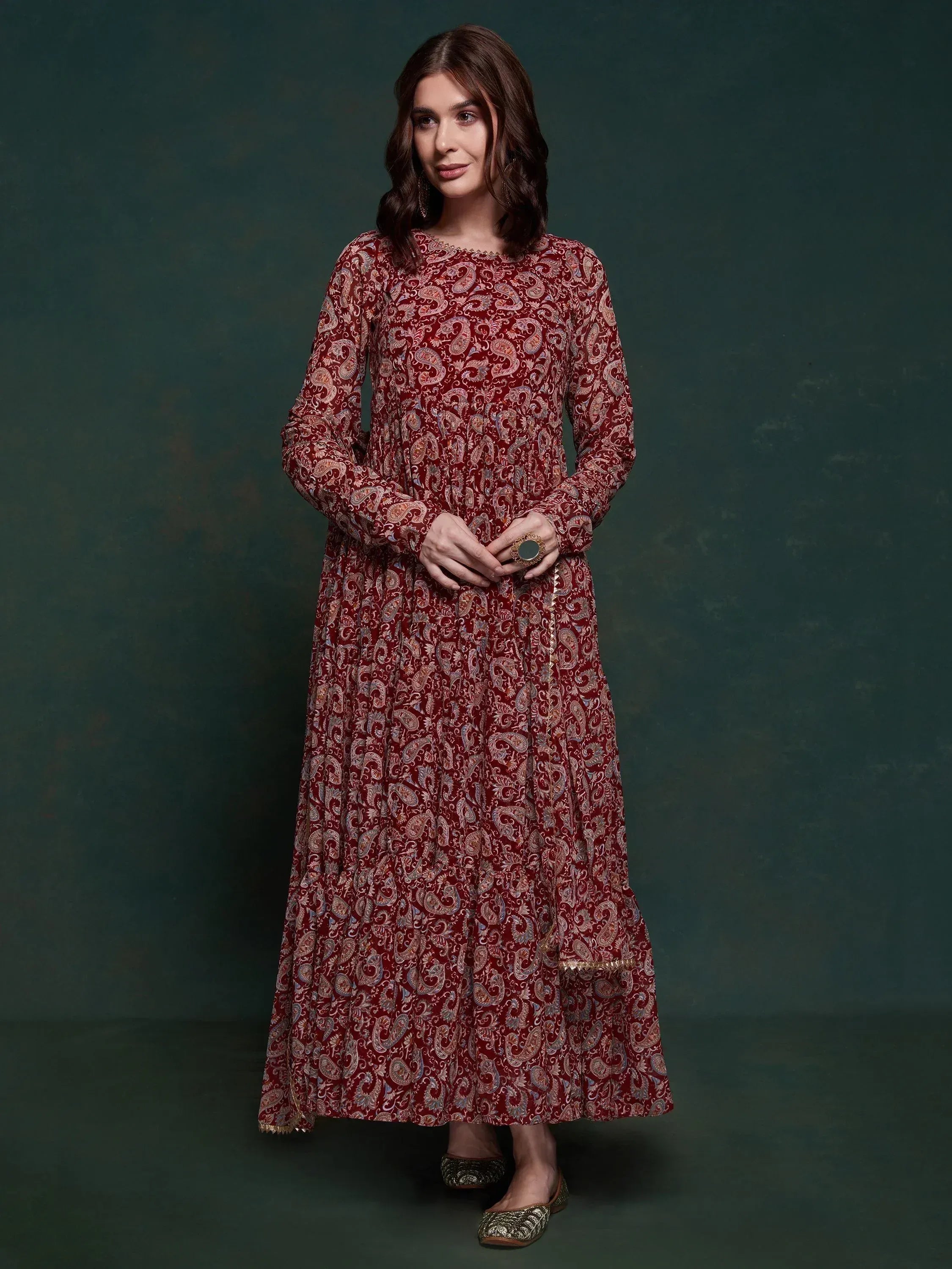 Woman in a maroon paisley print ethnic gown with long sleeves, standing against a dark background