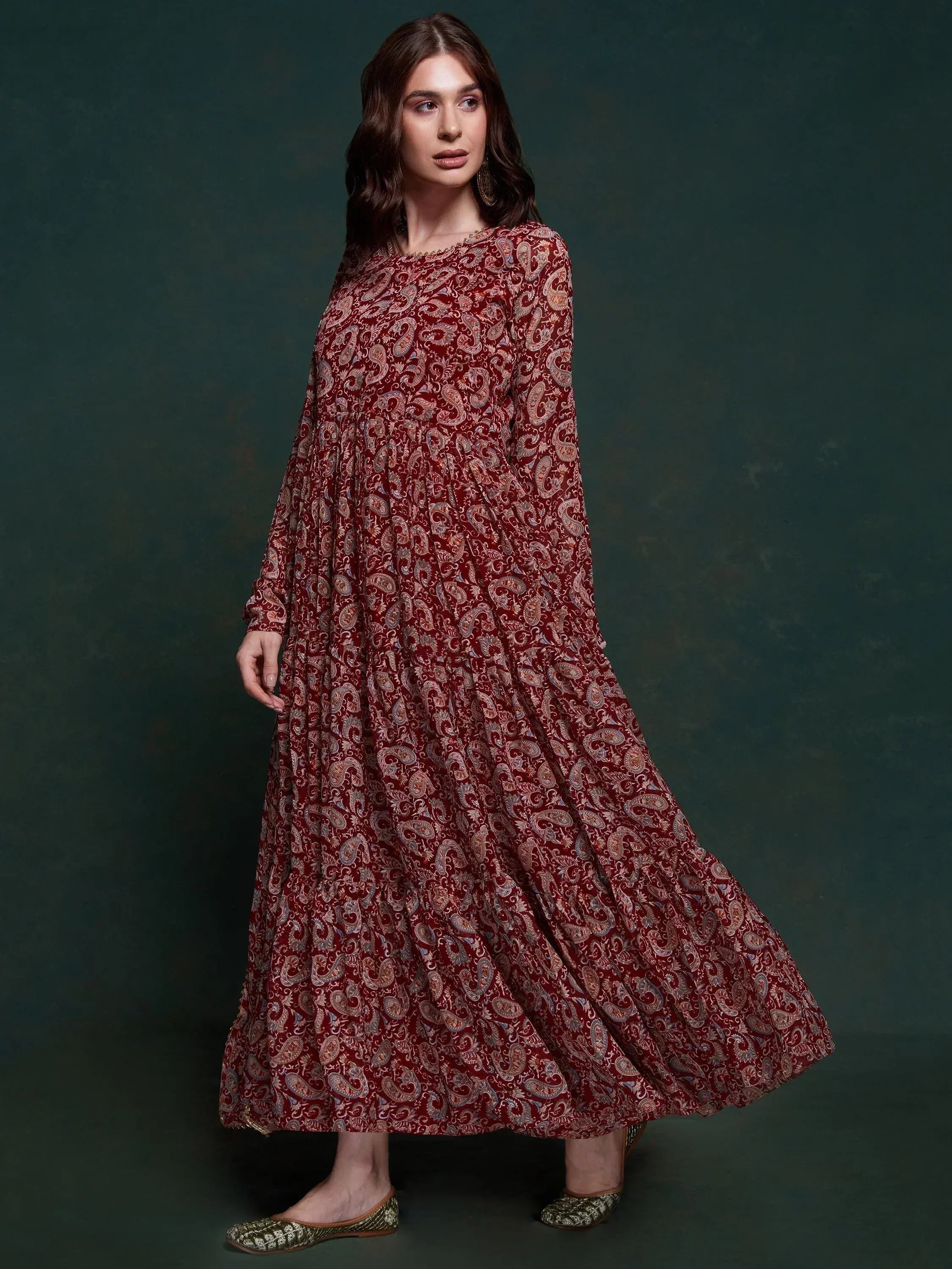 Woman wearing maroon paisley print ethnic gown with long sleeves, standing against dark background