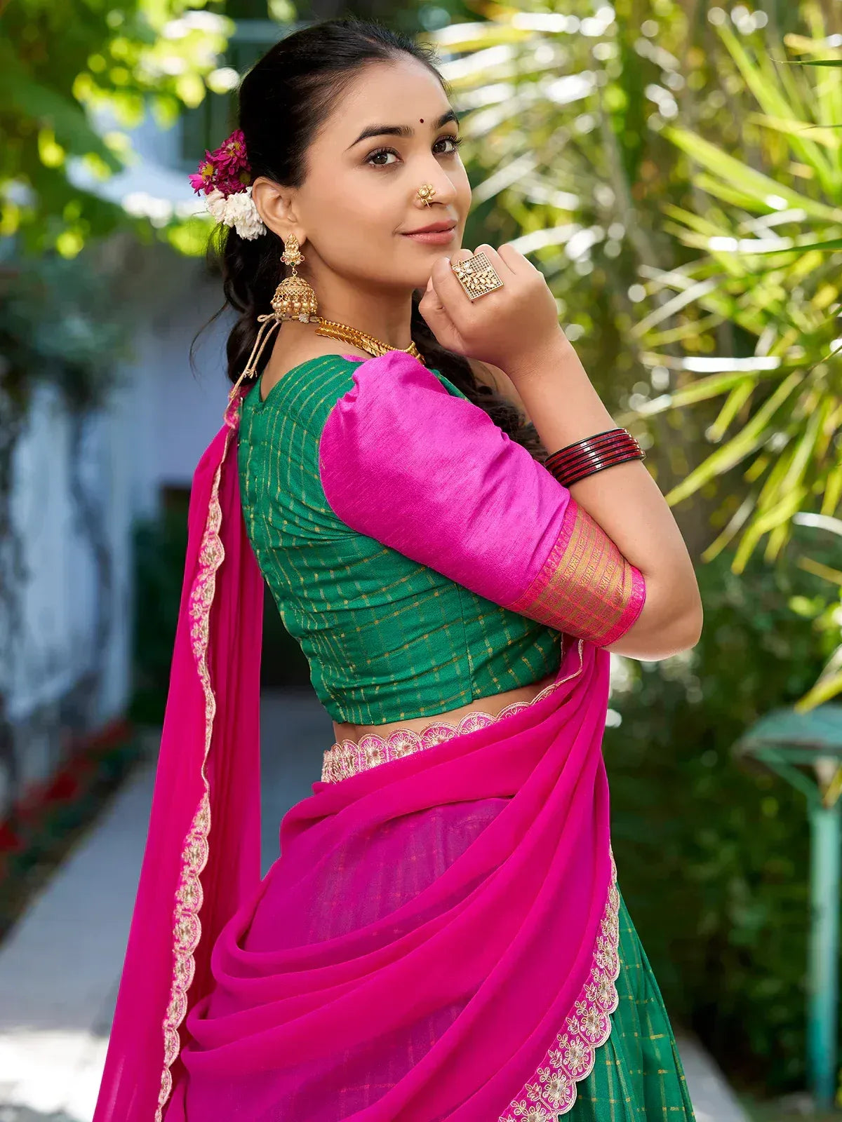 Woman in vibrant pink and green ethnic saree with gold embroidery, styled with traditional jewelry and floral hair accessory