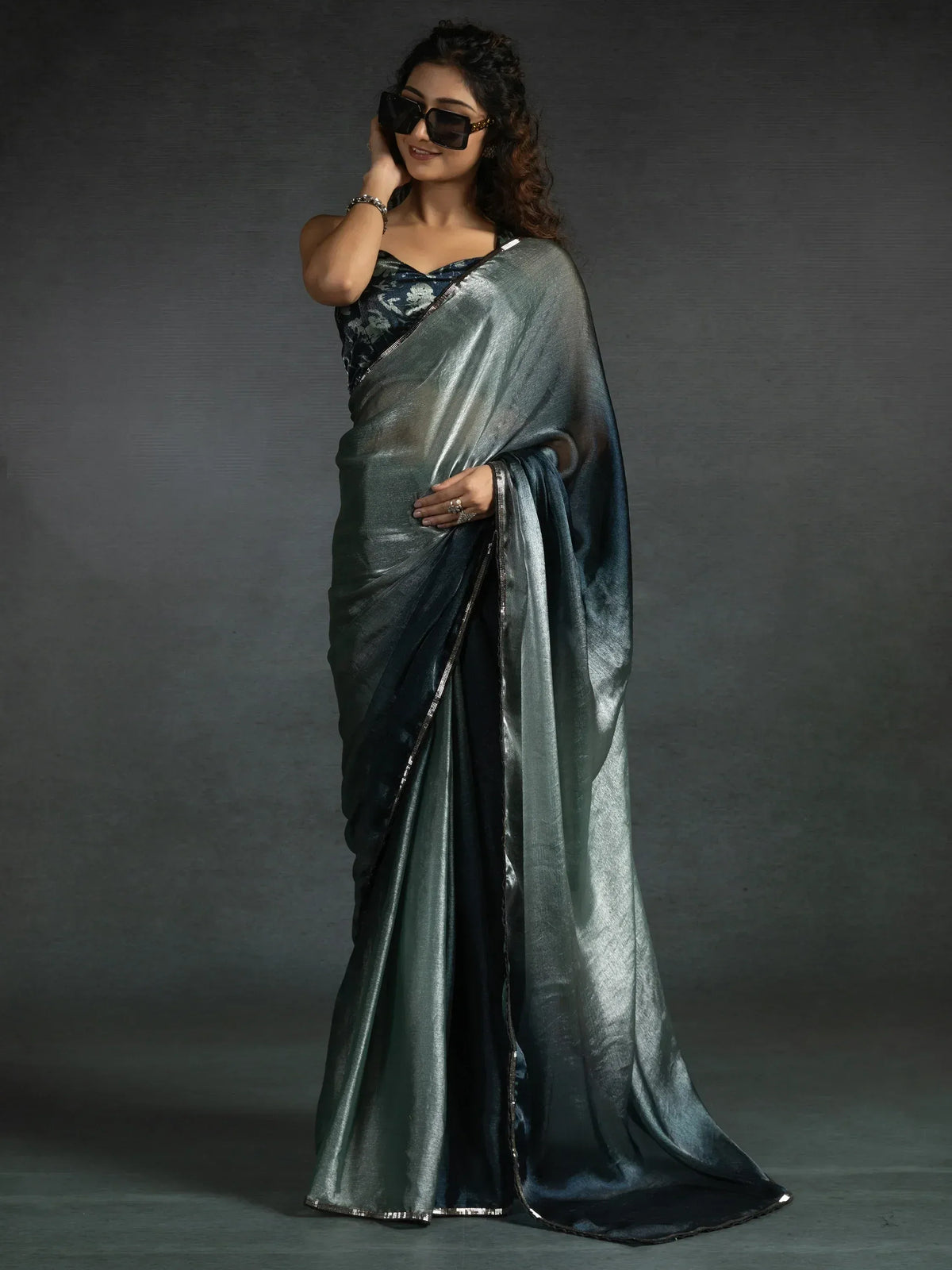 Woman in elegant dark green metallic saree with floral blouse, styled with sunglasses for a modern ethnic look.