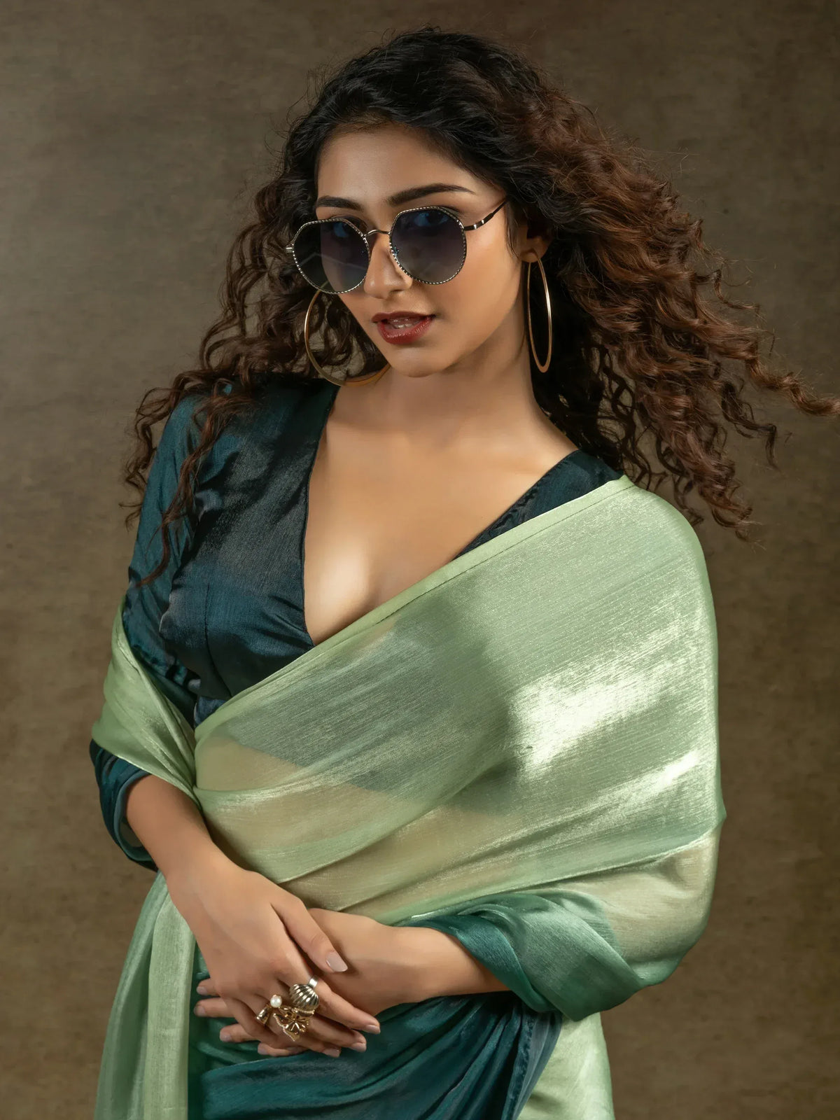 Stylish woman in green saree with deep neckline, sunglasses, and gold hoop earrings