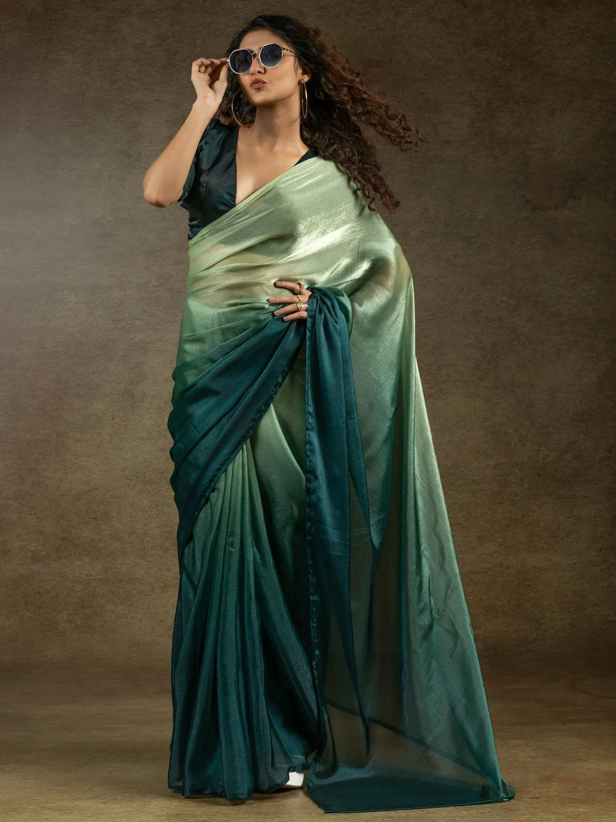 Woman in elegant green ombre saree with matching blouse, stylish sunglasses, and hoop earrings