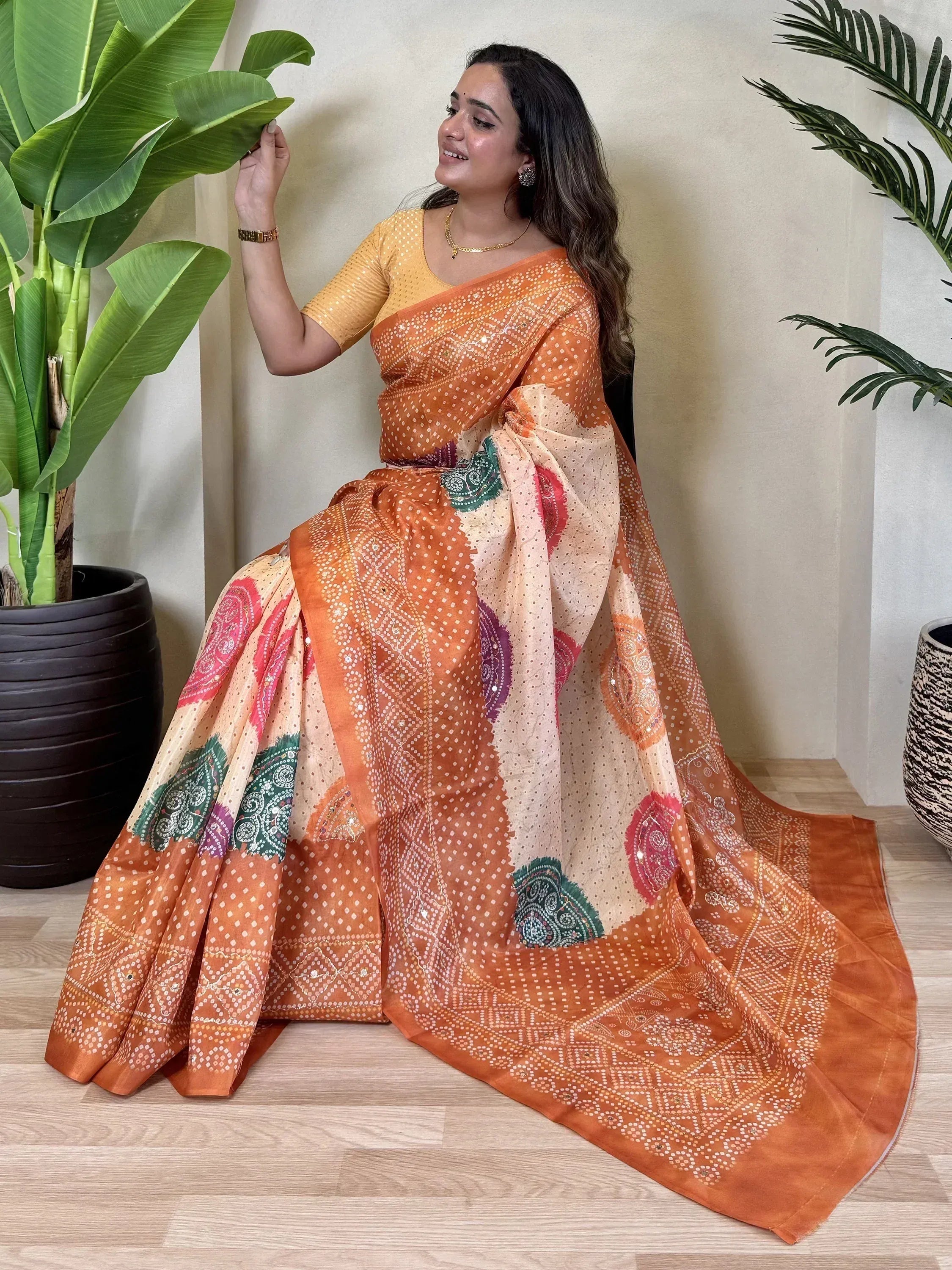 Woman in orange printed saree with yellow blouse, ethnic Indian wear, paisley and dot patterns, indoor plant backdrop