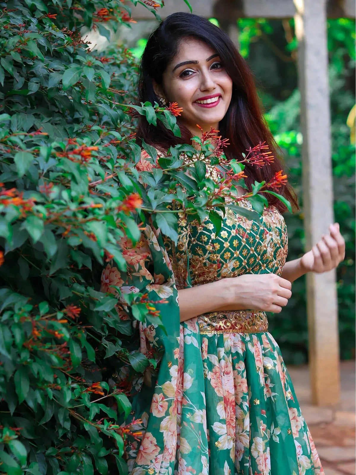 Woman in green floral ethnic gown with gold embroidery, smiling near garden foliage