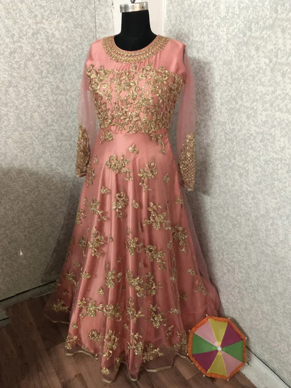 Pink ethnic gown with gold floral embroidery, long sleeves, displayed on mannequin with colorful umbrella