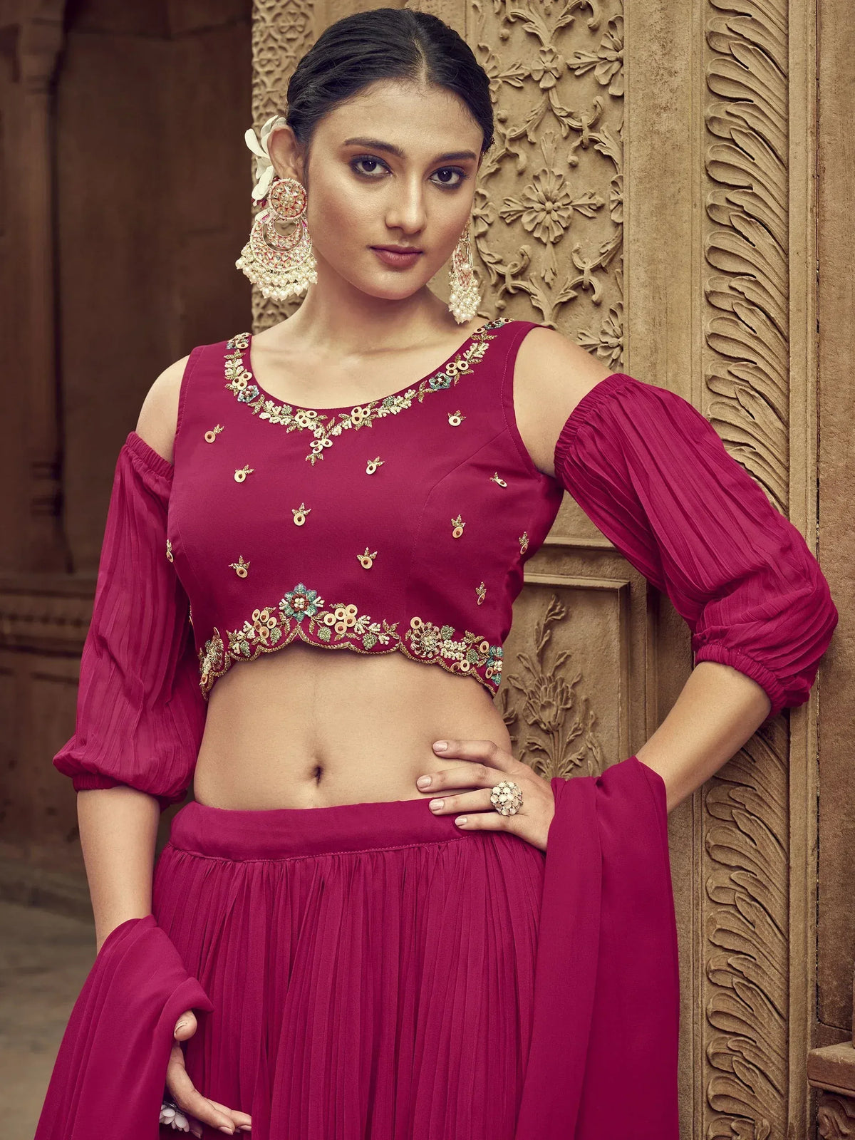 Woman in maroon embroidered lehenga choli with cold shoulder top, floral jewelry, against carved wall
