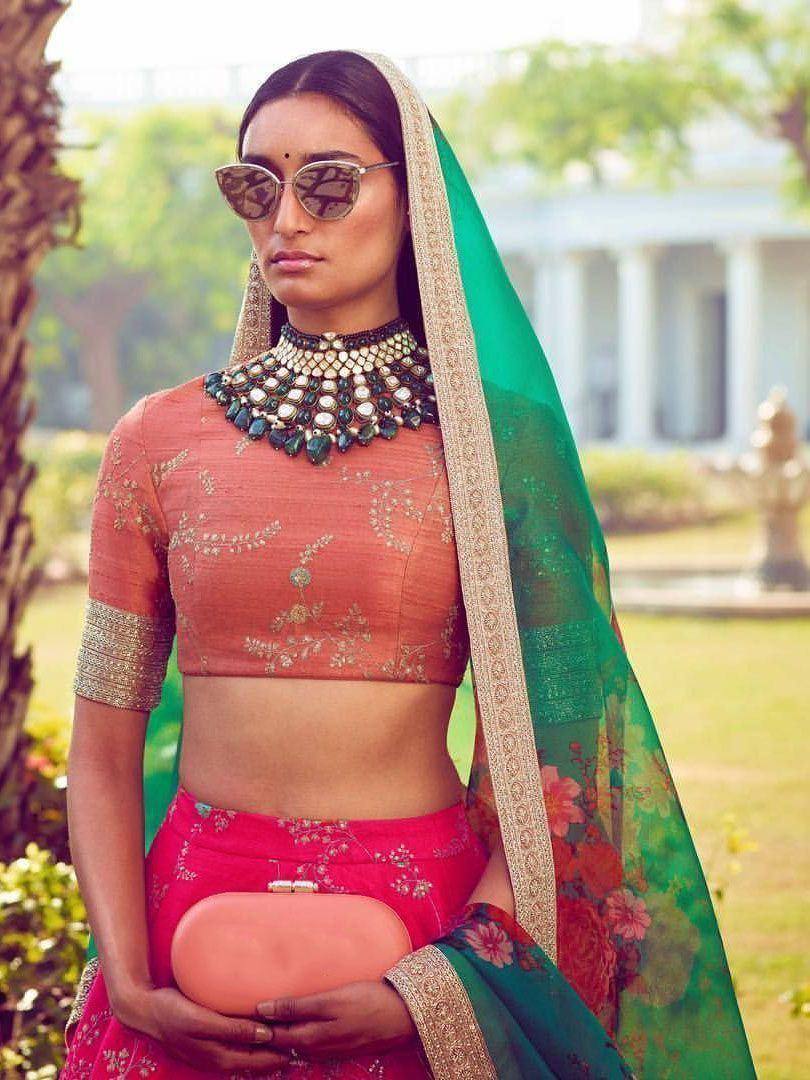 Woman in pink embroidered lehenga choli with green floral dupatta, statement necklace, and sunglasses