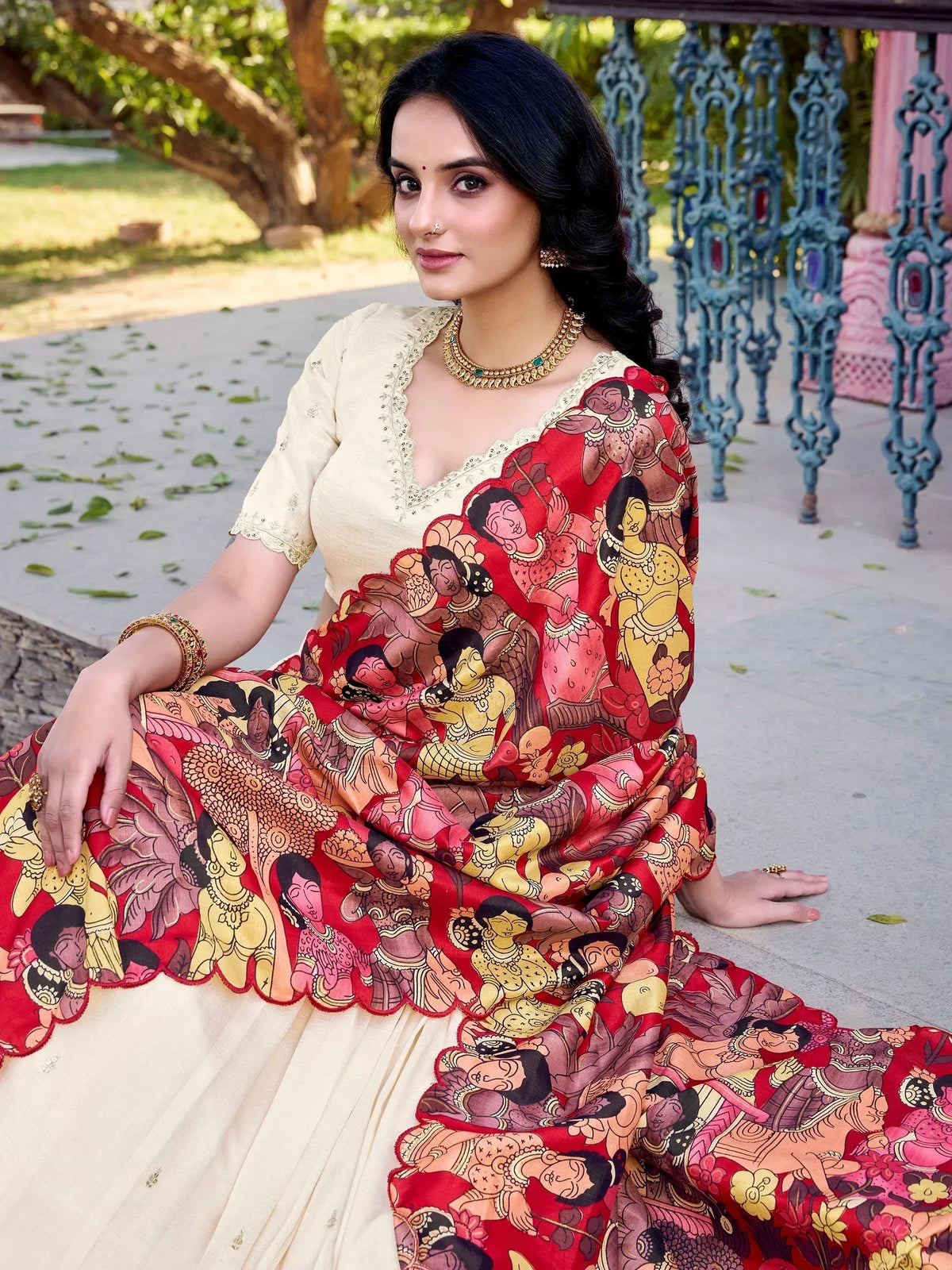 Woman in red printed saree with ethnic artwork, cream blouse, traditional jewelry, outdoor setting