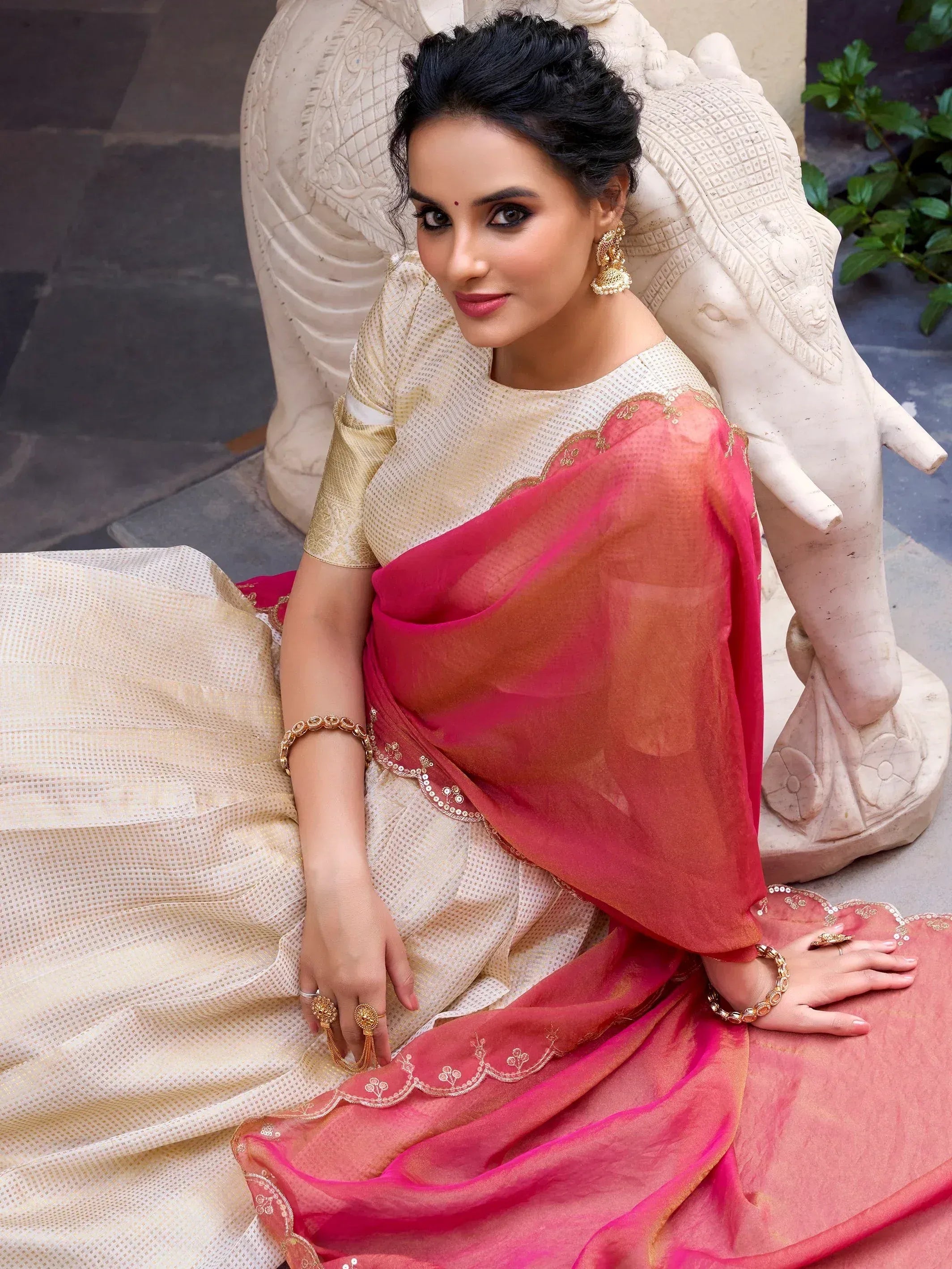 Woman in elegant cream and pink saree with gold embroidery, styled with traditional jewelry, seated beside stone elephant sculpture