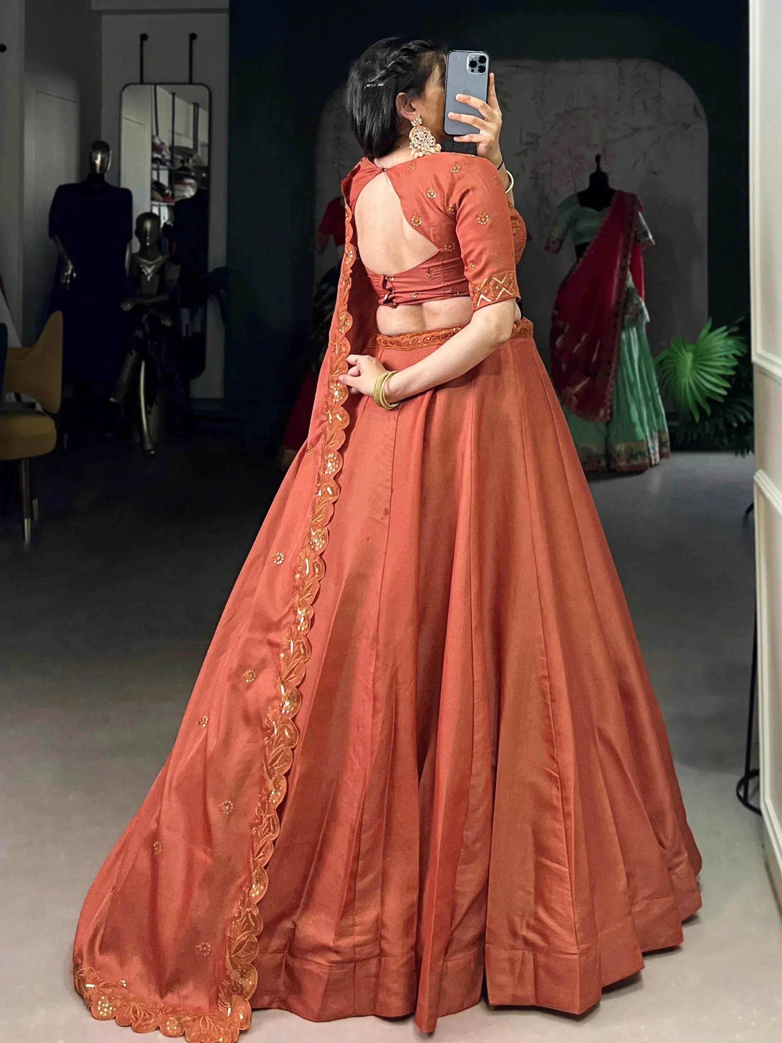 Woman in rust orange designer lehenga choli with backless blouse and gold embroidery, taking mirror selfie