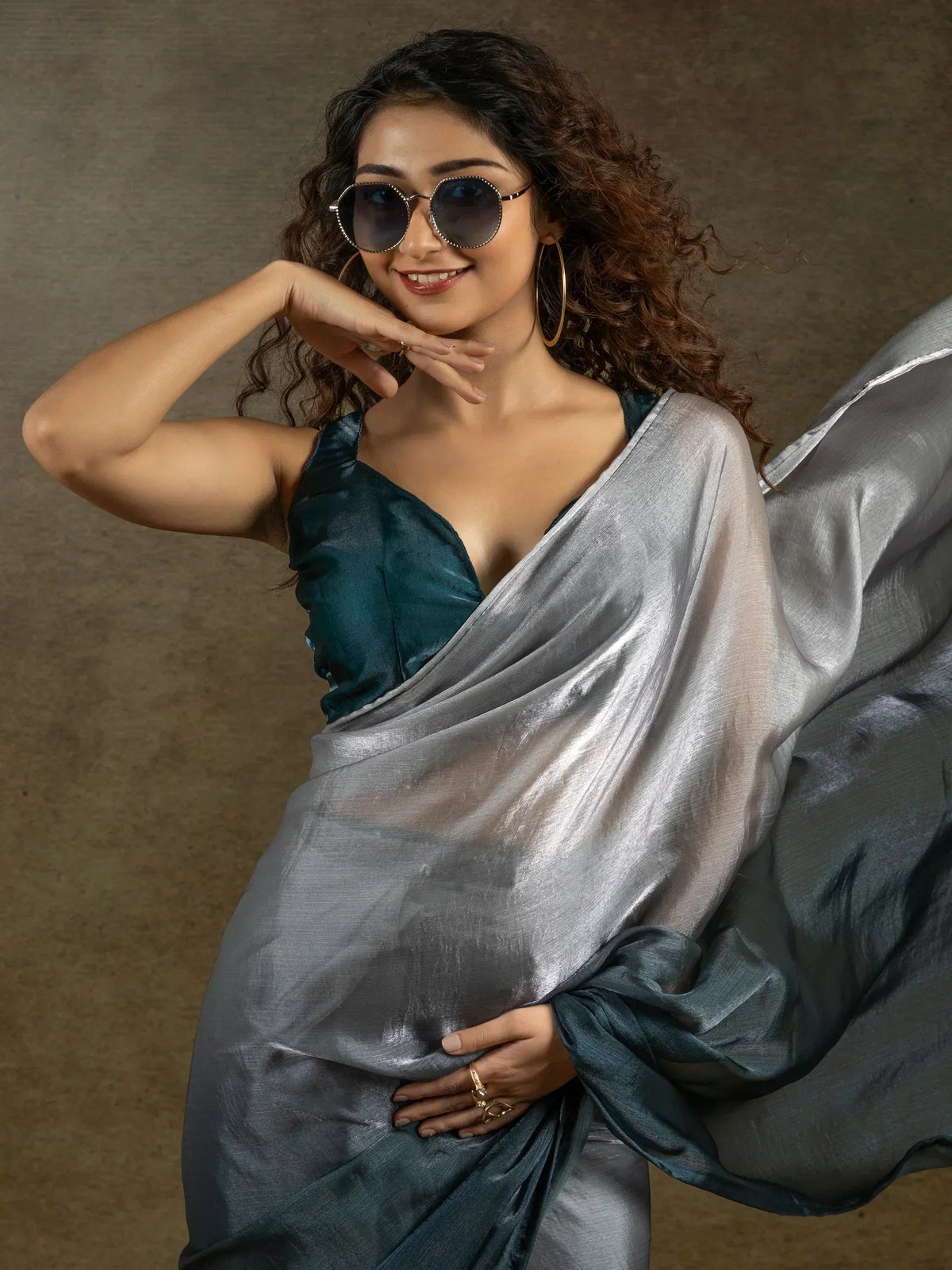 Stylish woman in silver saree with teal blouse, wearing sunglasses and gold hoops, posing confidently.