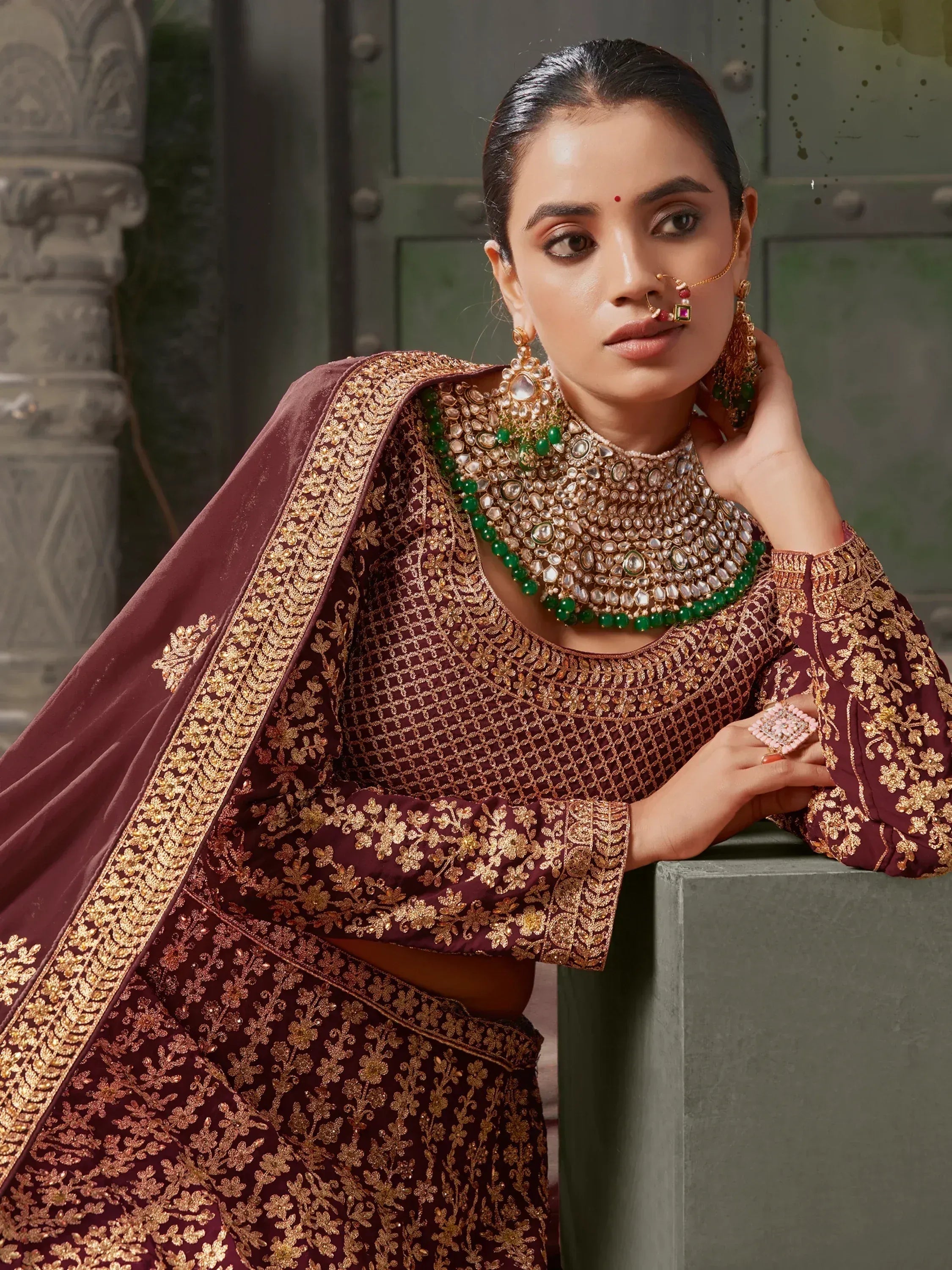 Maroon embroidered lehenga choli with gold work, green jewelry, and traditional Indian bridal accessories
