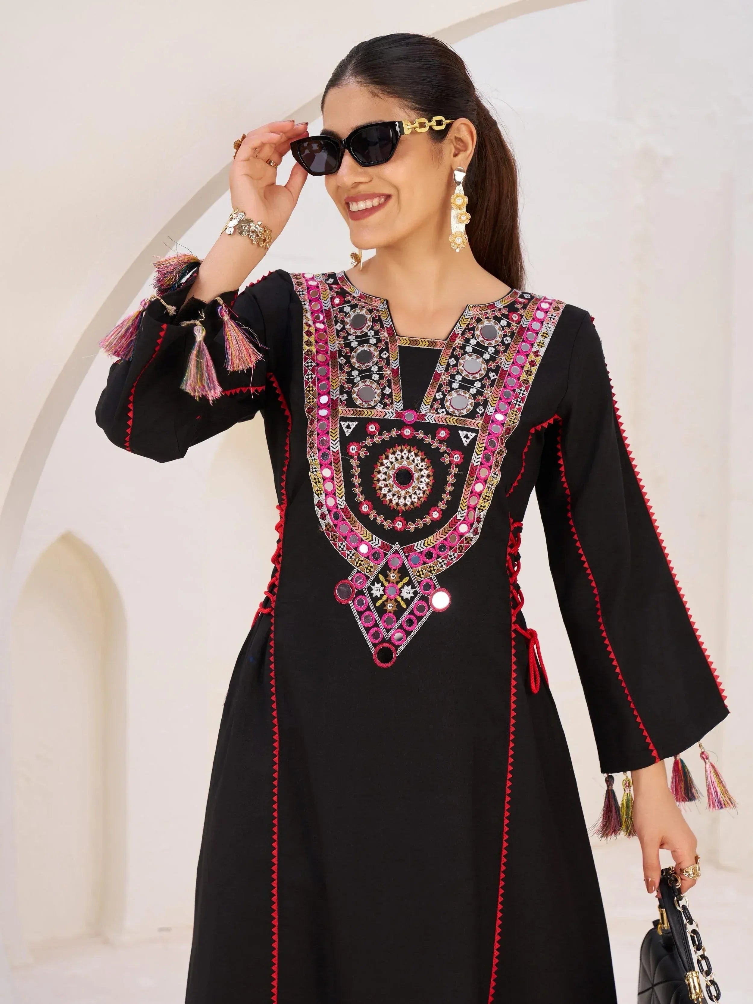 Black ethnic kurta with mirror work, tassels, and pink embroidery, styled with sunglasses and gold jewelry.