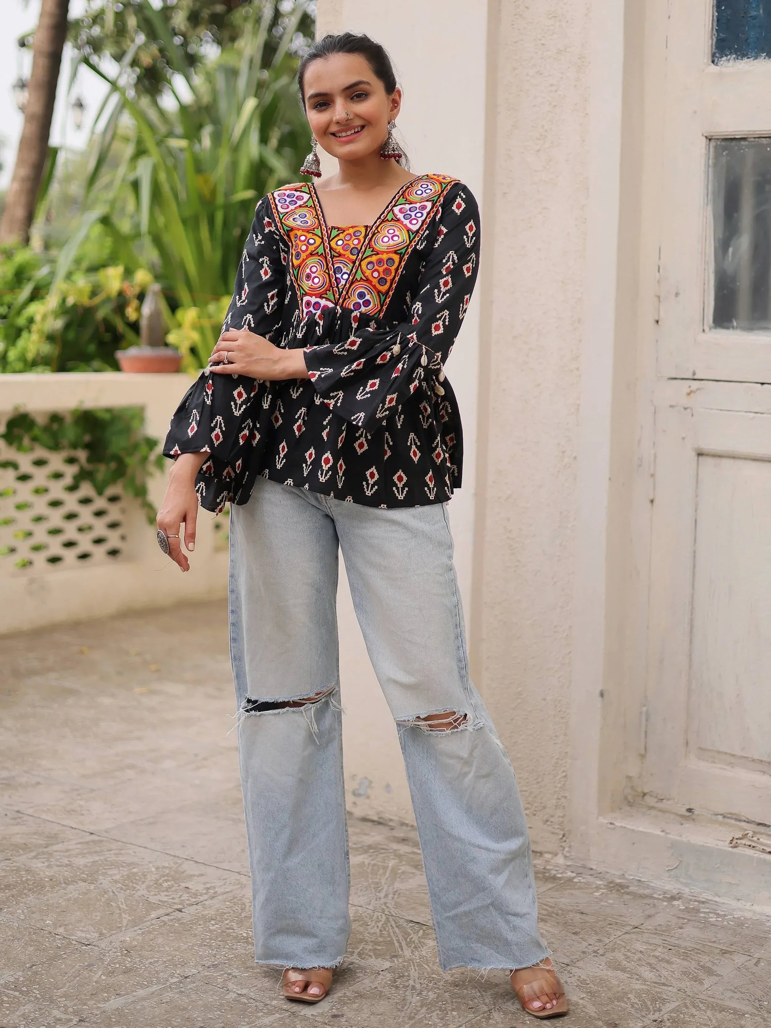 Woman in black embroidered ethnic top with colorful patterns, paired with ripped wide-leg jeans and jhumka earrings.
