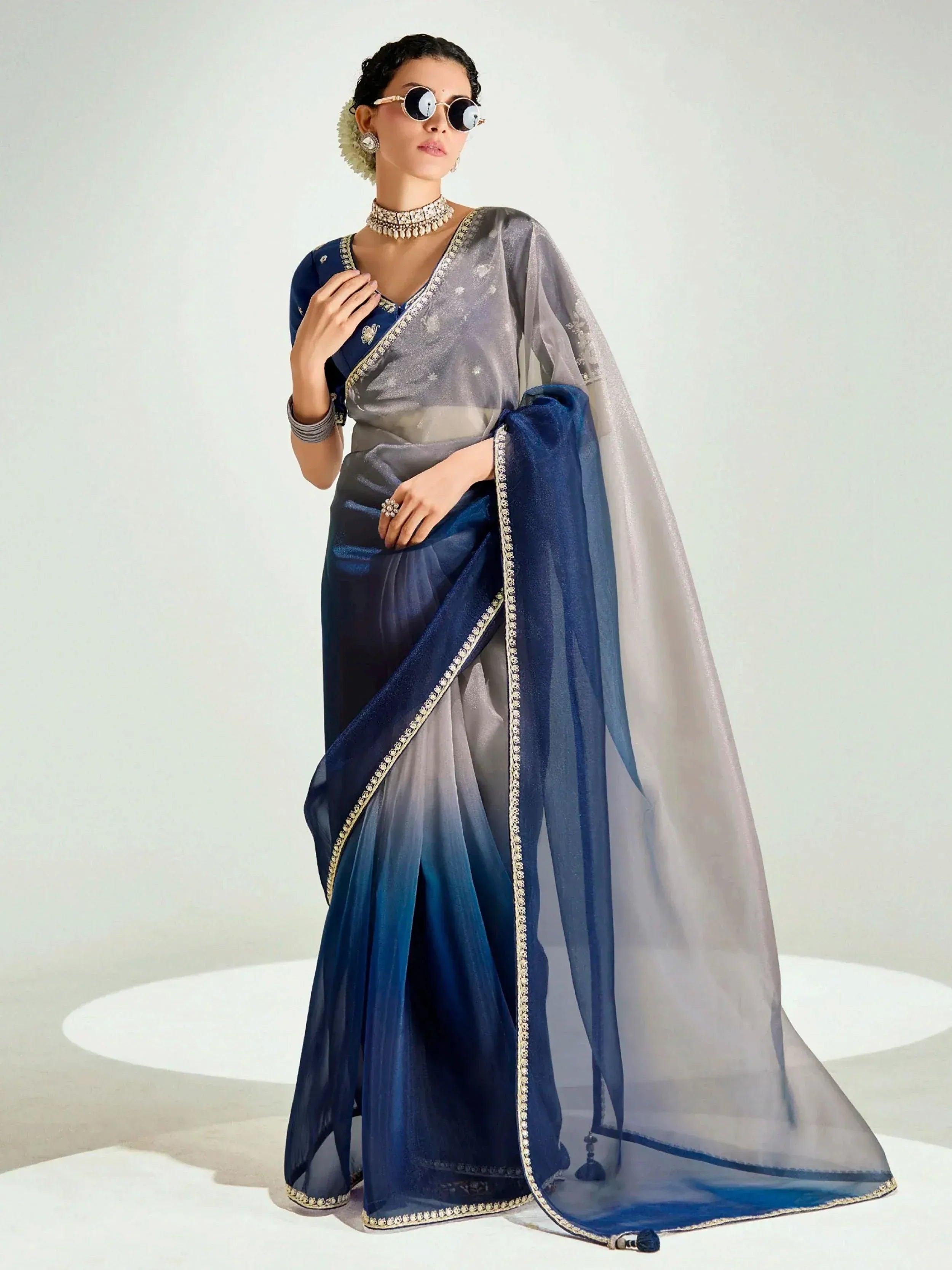 Elegant blue and gray ombre saree with gold embroidery, styled with traditional jewelry and sunglasses.