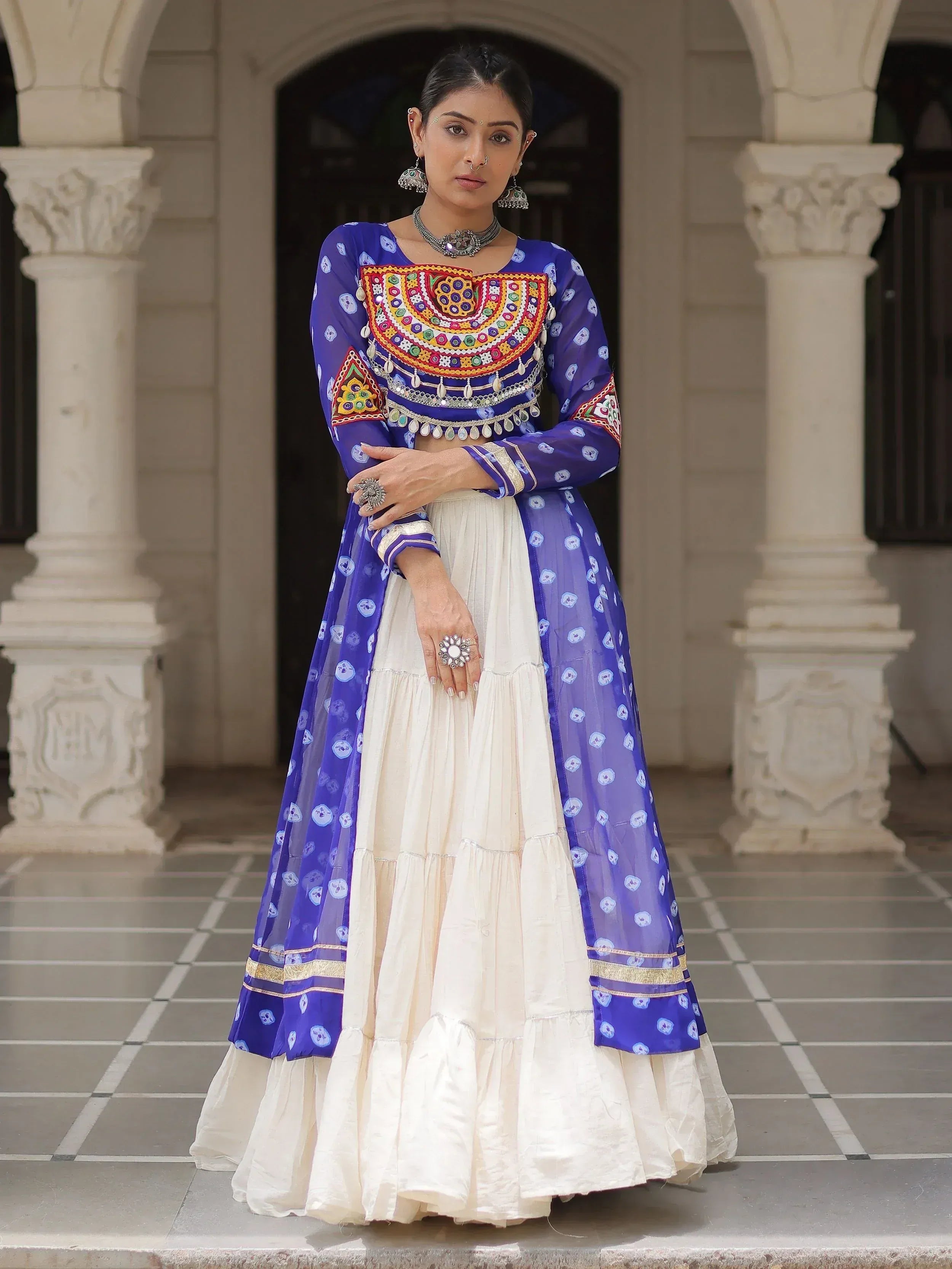 Woman in blue embroidered Indian ethnic wear with white lehenga, traditional jewelry, standing at arched entrance