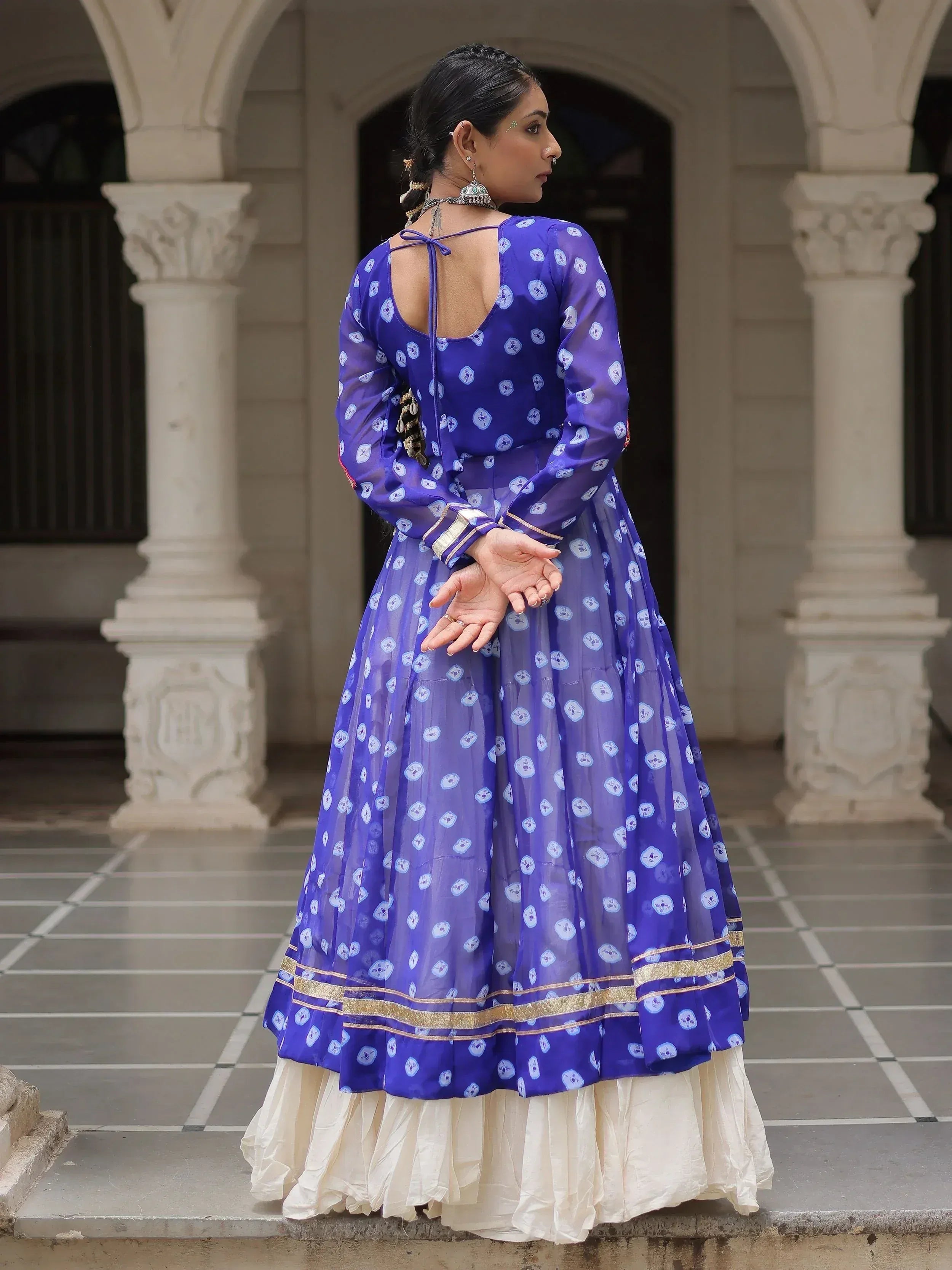 Woman in purple printed ethnic gown with gold trim, back view, standing in arched courtyard