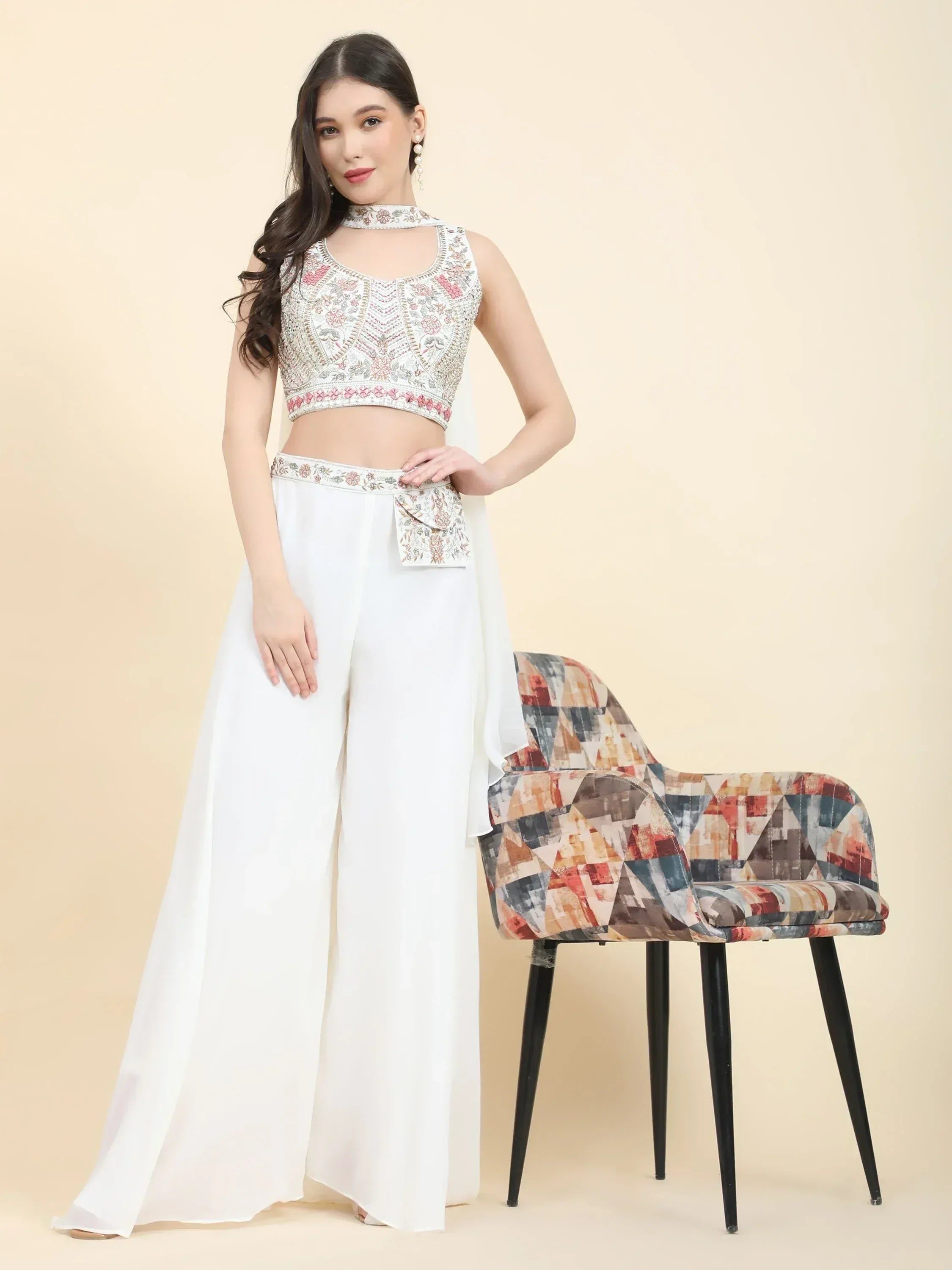 Woman in white embroidered crop top and palazzo pants, posing by a patterned chair