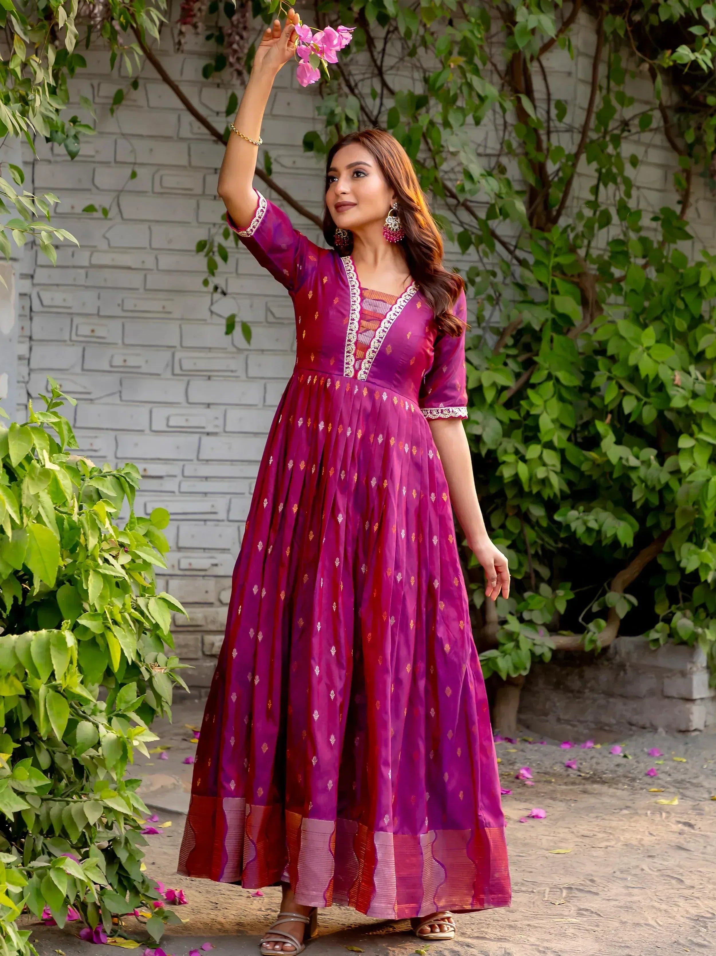 Woman in purple ethnic gown with gold embroidery, holding pink flowers against green foliage