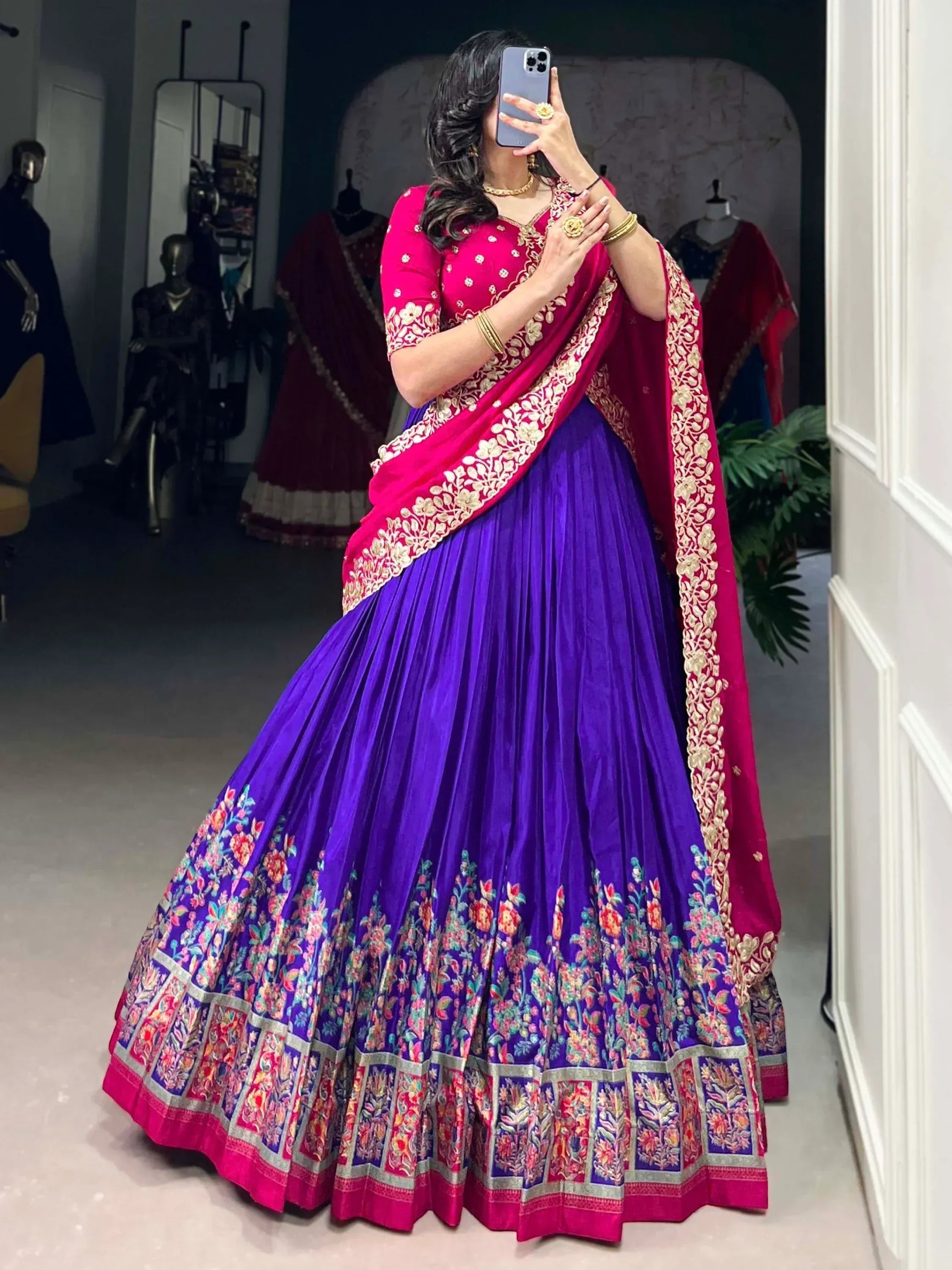 Woman in vibrant purple and pink embroidered lehenga choli with floral border, taking mirror selfie in boutique.