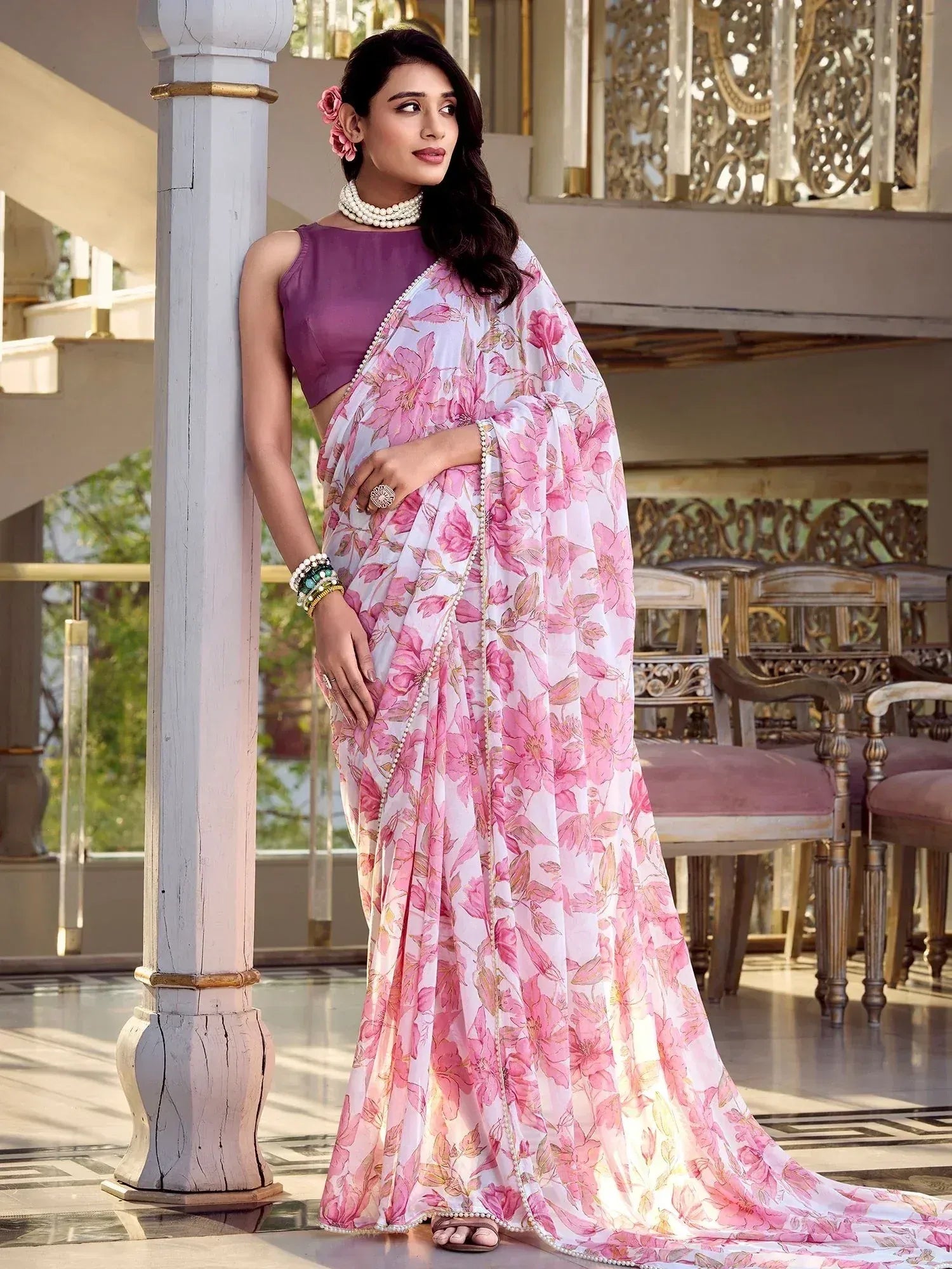 Woman in pink floral saree with purple blouse, pearl jewelry, leaning on pillar