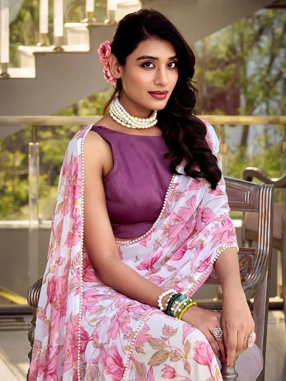 Woman in purple sleeveless top with pink floral saree, pearl jewelry, and hair flowers, seated outdoors
