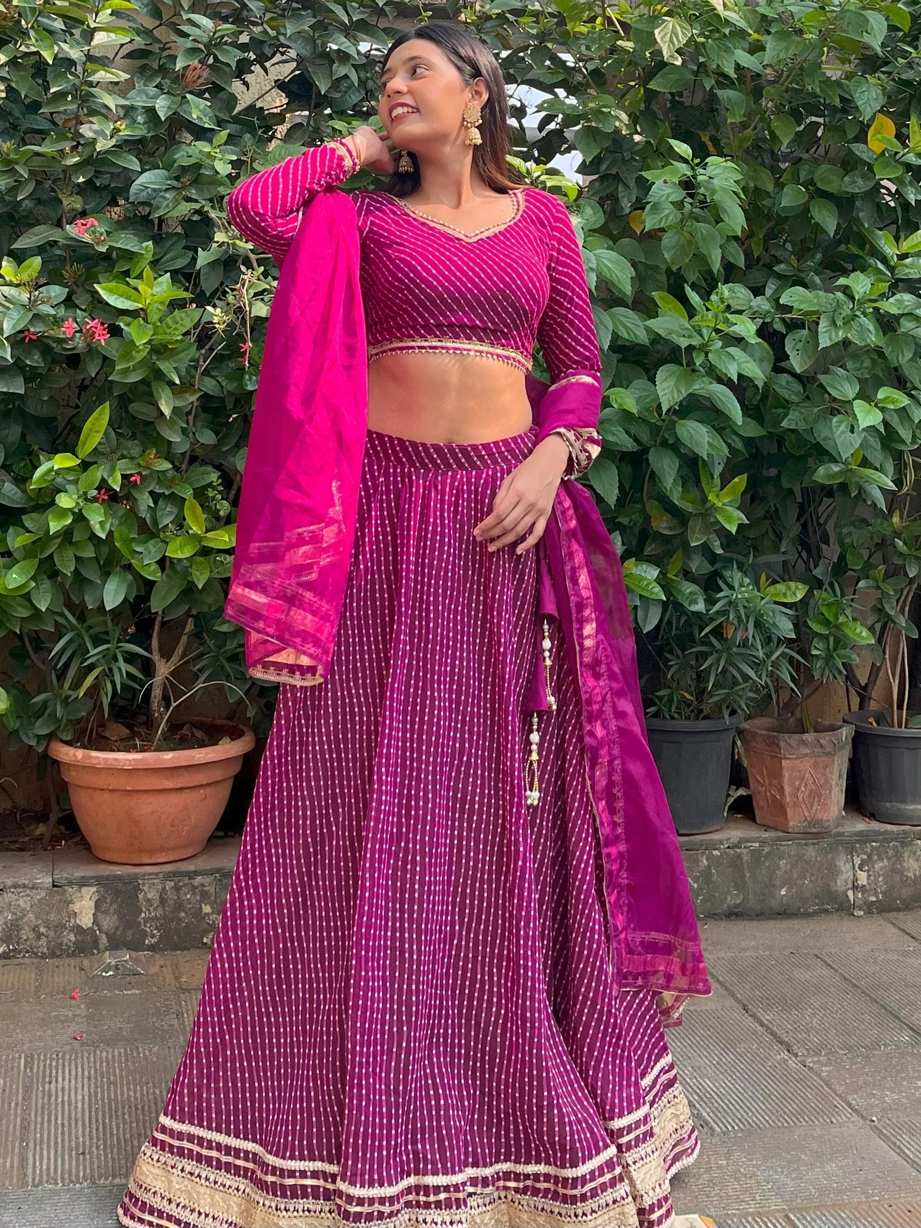 Woman in vibrant pink and purple striped lehenga choli with gold trim, standing outdoors near greenery