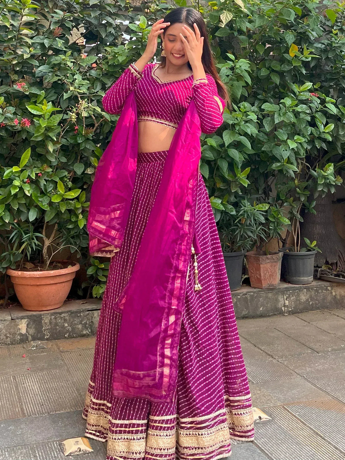 Woman in vibrant pink striped lehenga choli with gold trim, posing outdoors near greenery