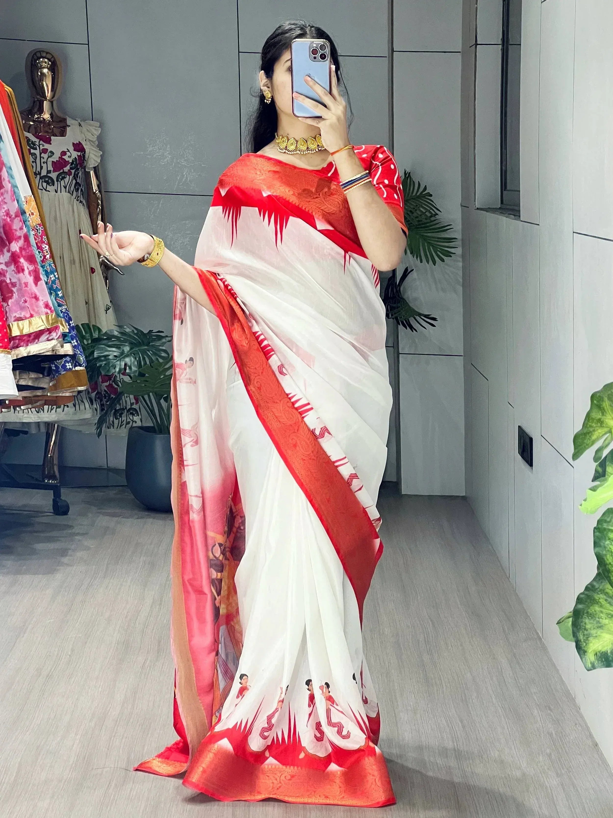Woman in white and red printed saree with gold jewelry, taking mirror selfie in ethnic wear store