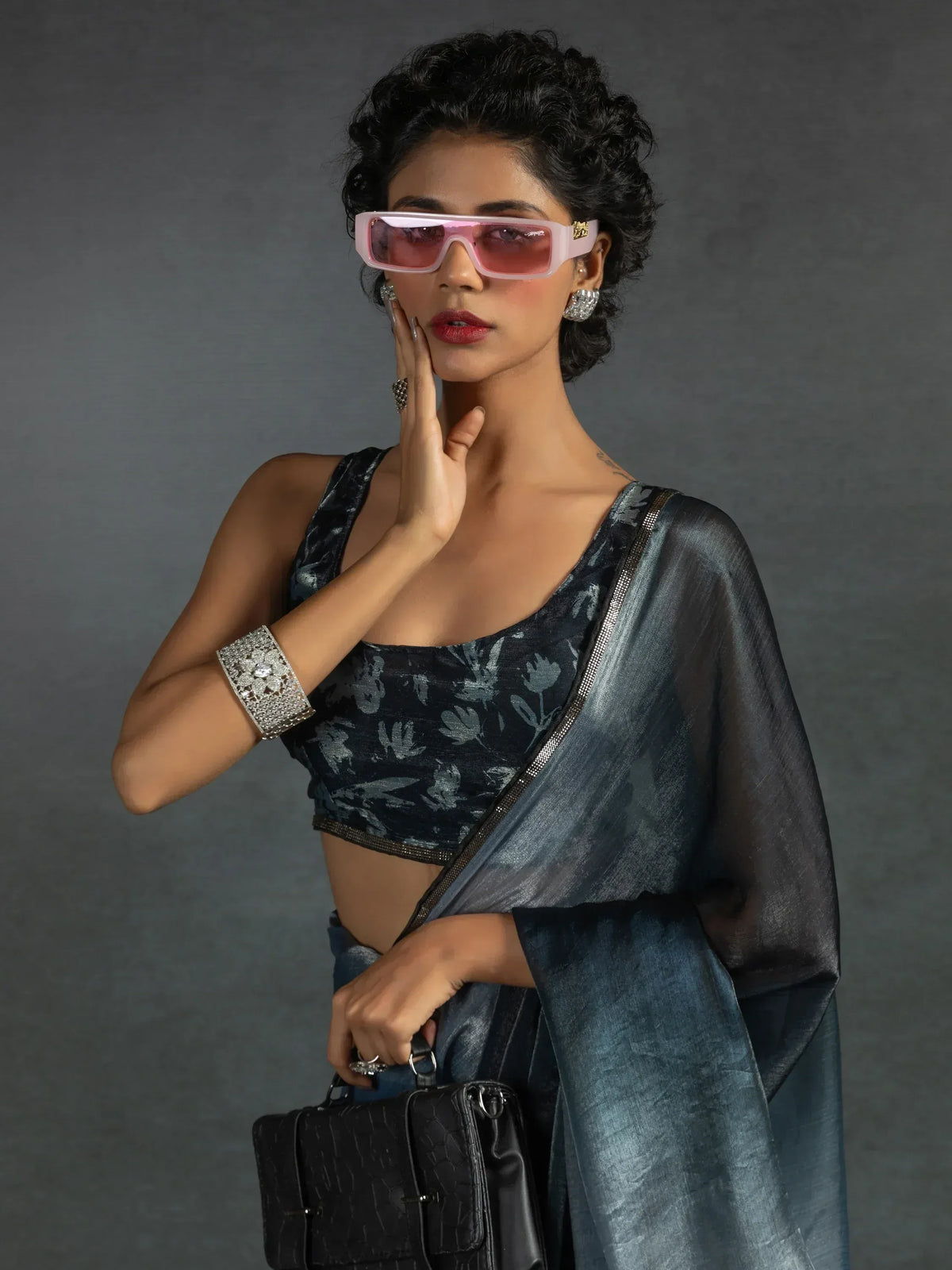 Stylish woman in blue floral saree with pink sunglasses and statement jewelry, holding black handbag
