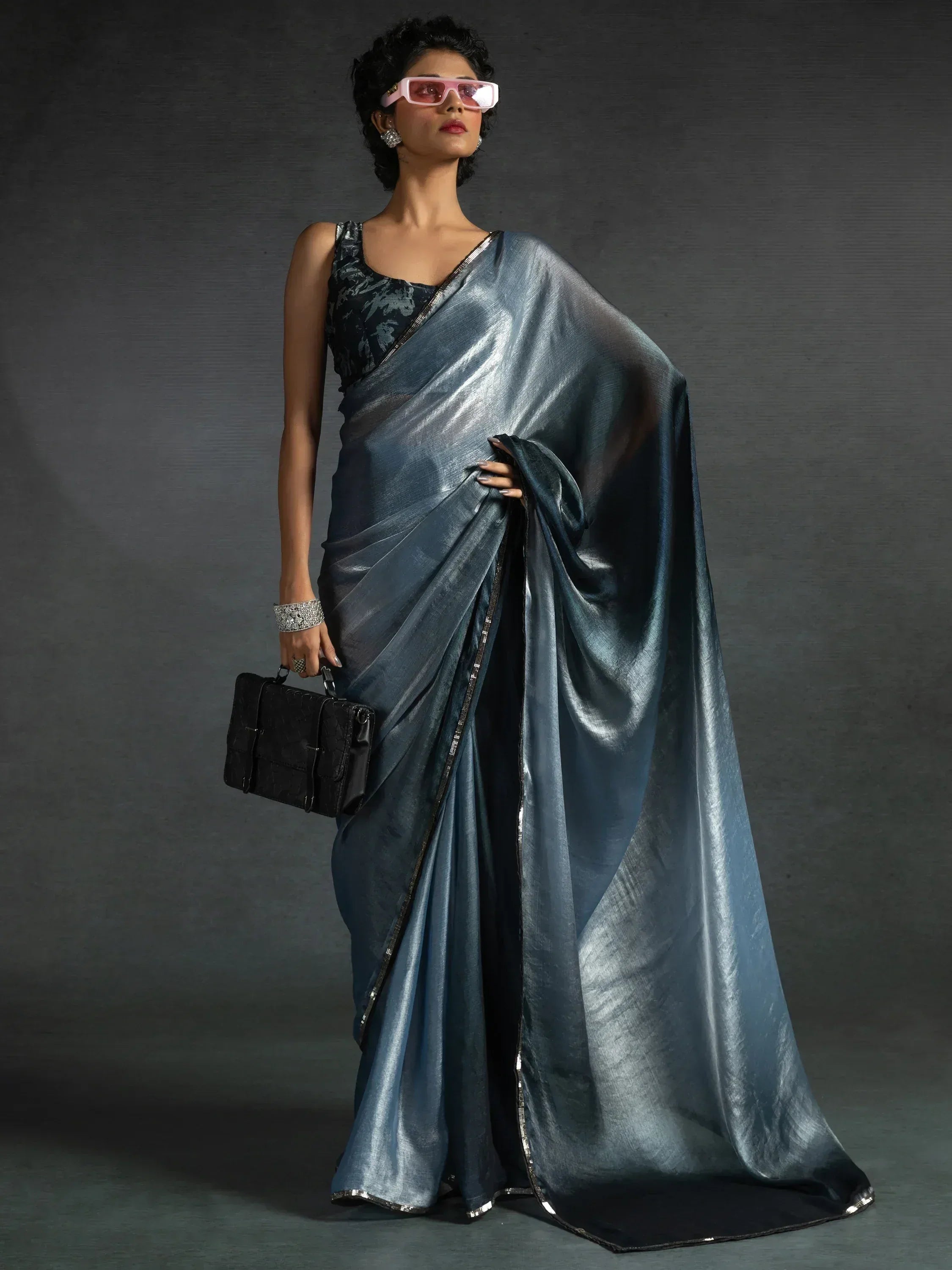 Stylish woman in metallic blue saree with black blouse, pink sunglasses, and clutch bag