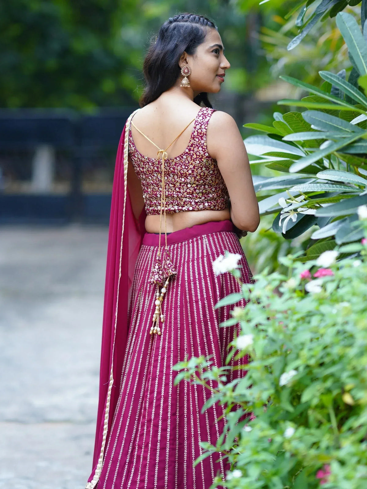 Woman in maroon designer lehenga choli with gold embroidery, back view in garden setting