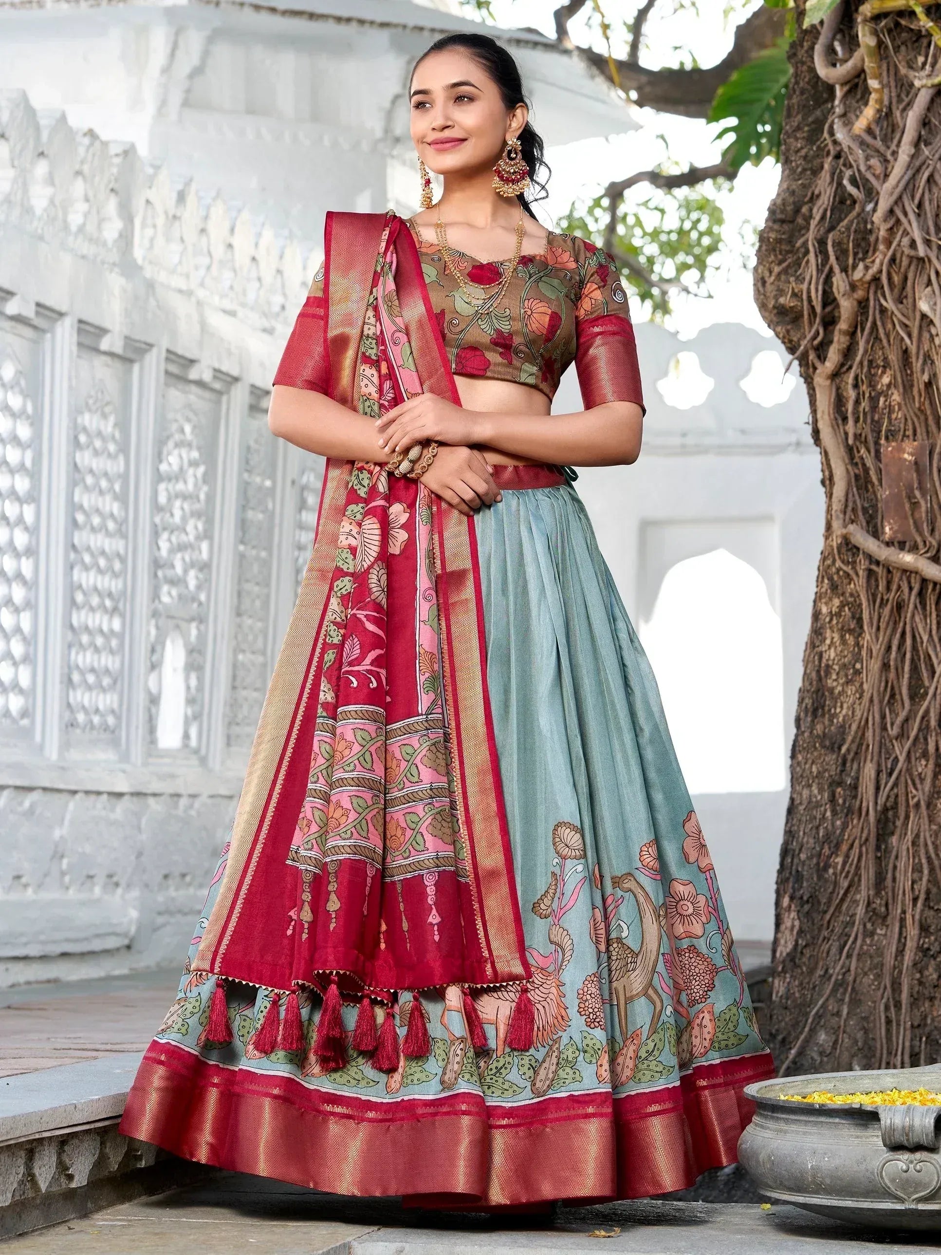Woman in designer lehenga choli with floral print, red dupatta, and tassels, standing near white architecture and tree