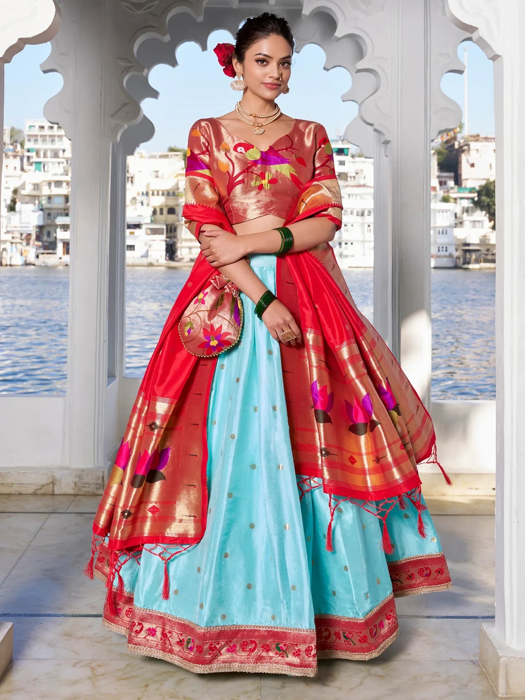 Woman in vibrant Indian lehenga choli with floral embroidery, standing by waterfront under ornate archway