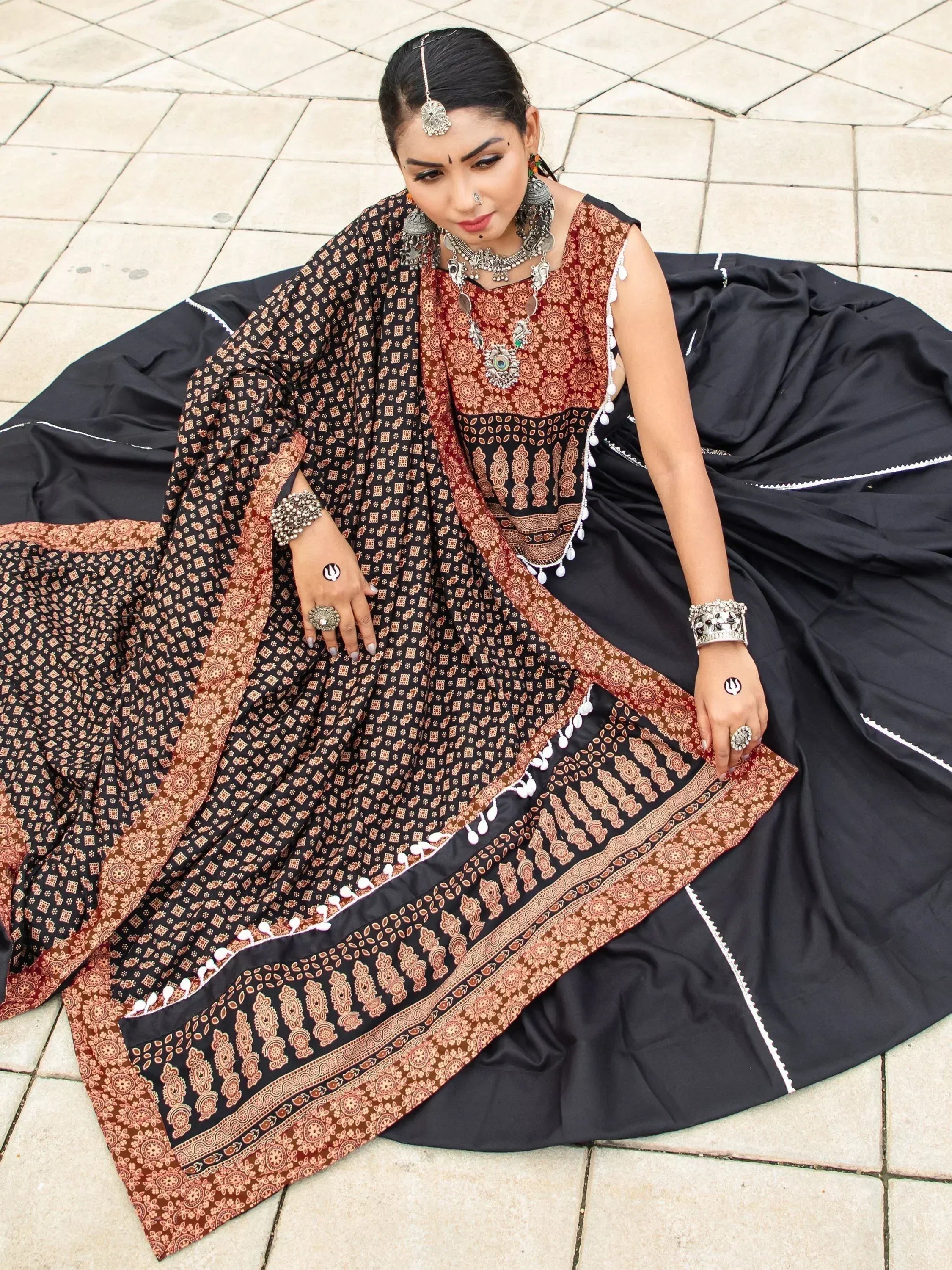 Woman in black and rust printed ethnic gown with mirror work, paired with traditional jewelry and dupatta.