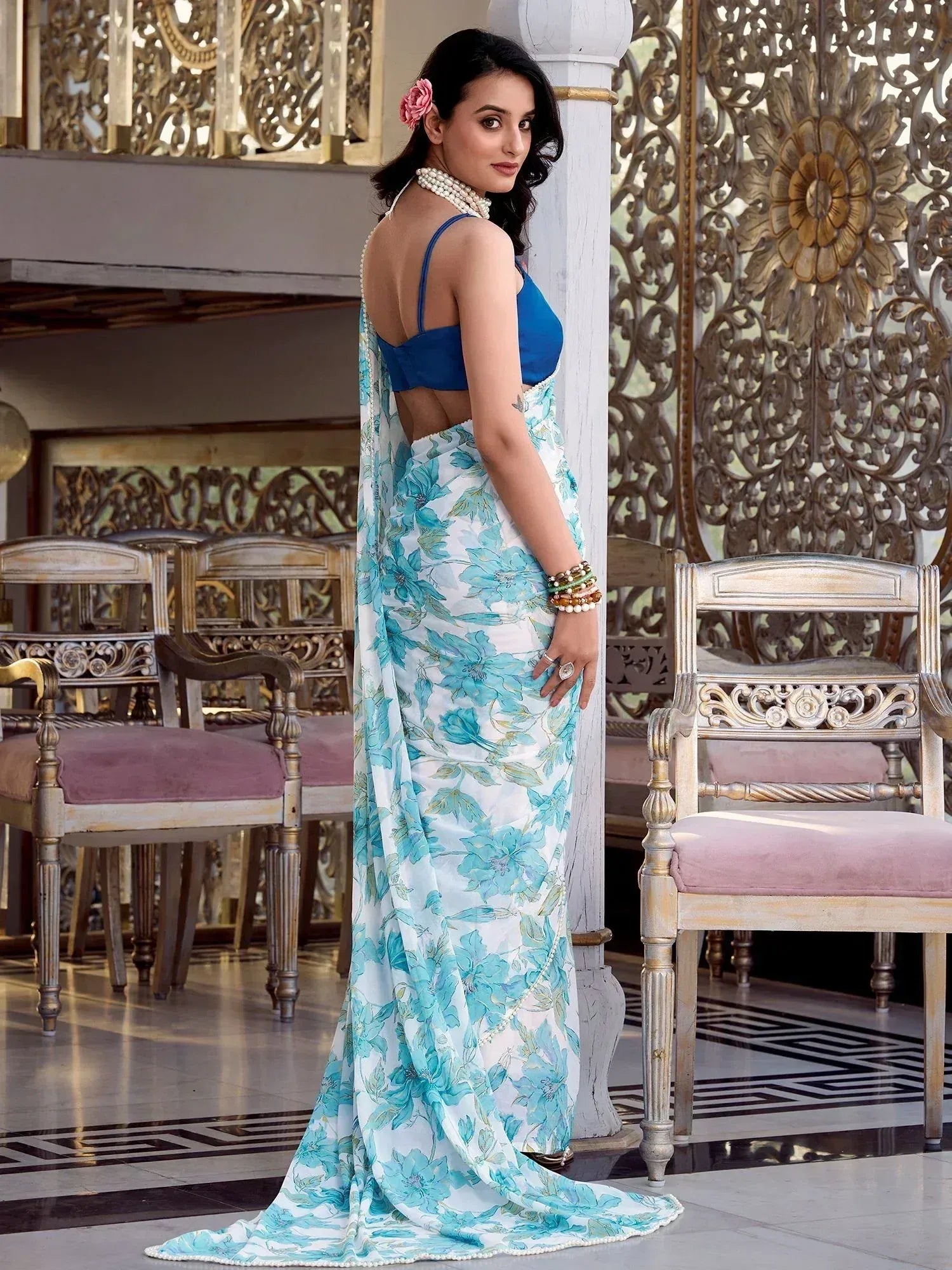 Woman in blue floral saree with backless blouse, pearl jewelry, and pink flower hair accessory