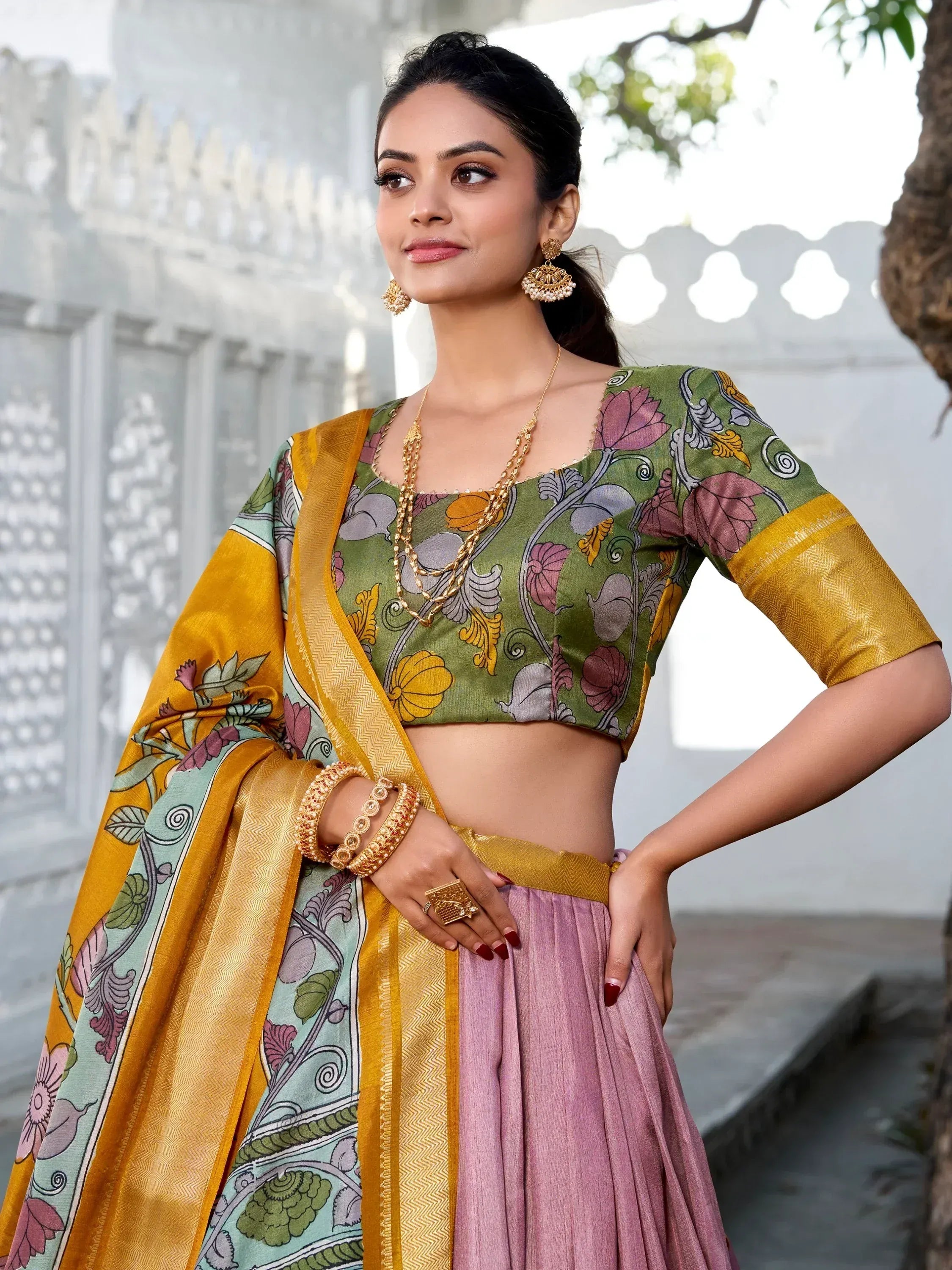 Woman in floral green blouse, pink lehenga, and yellow dupatta, styled with gold jewelry for ethnic wear.