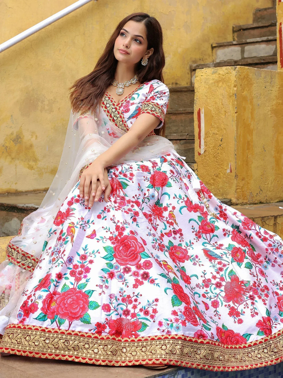 Woman in white floral lehenga choli with red roses, birds, and gold embroidery, sitting on steps