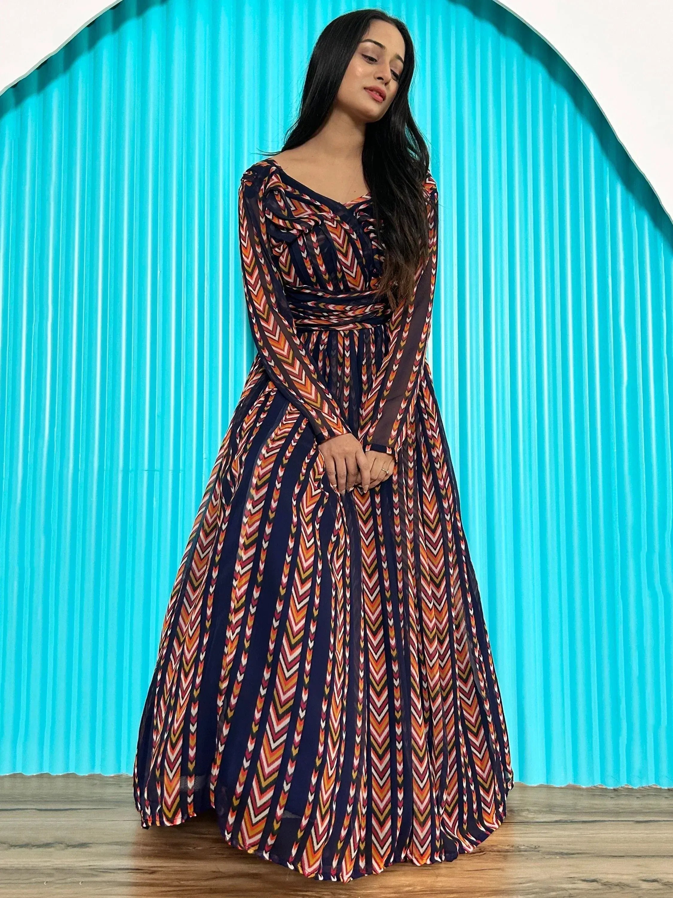 Woman in colorful ethnic gown with chevron print, long sleeves, and pleated skirt against blue backdrop
