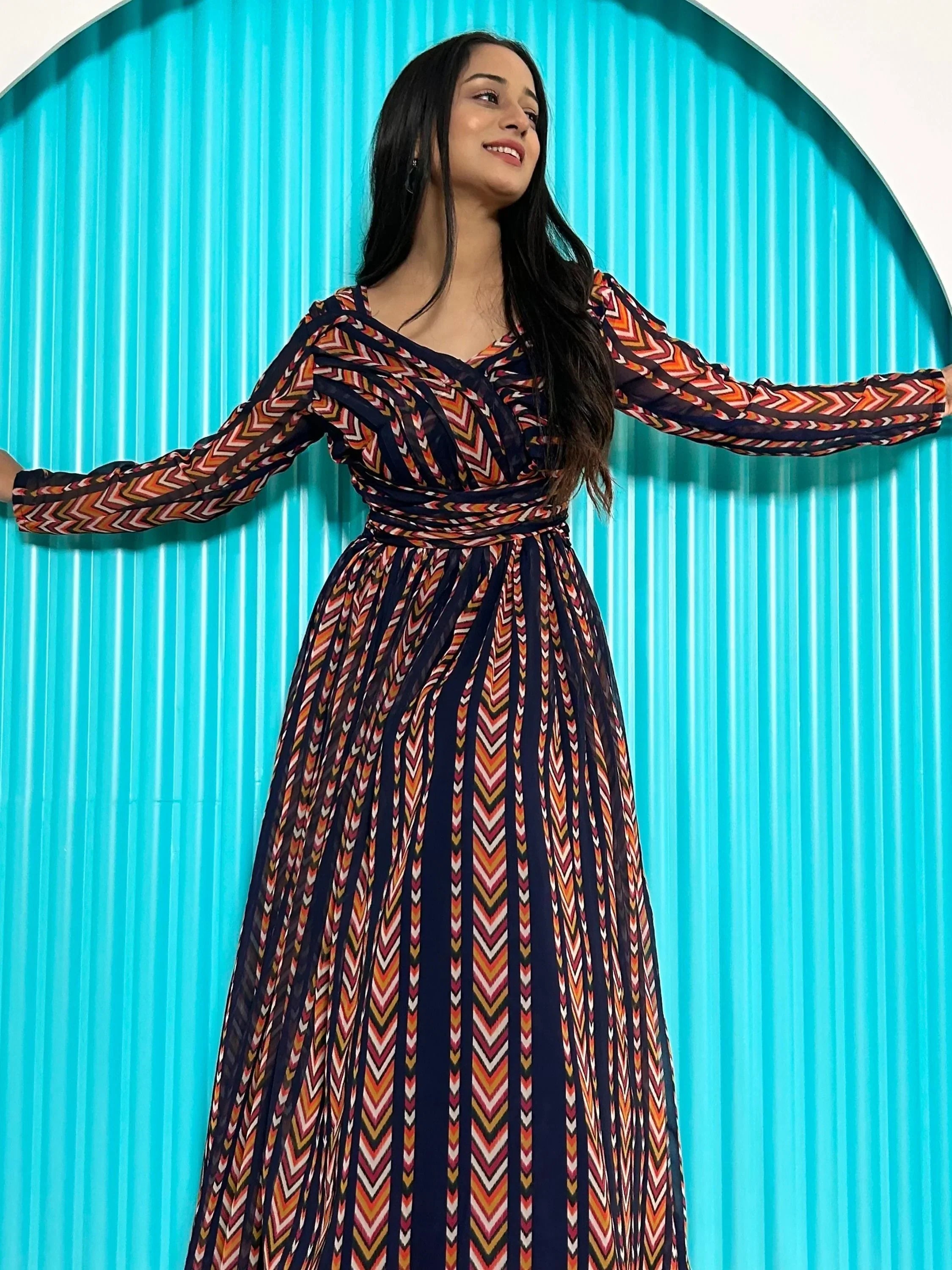 Woman in a long sleeve multicolor printed ethnic gown posing against a blue arched background