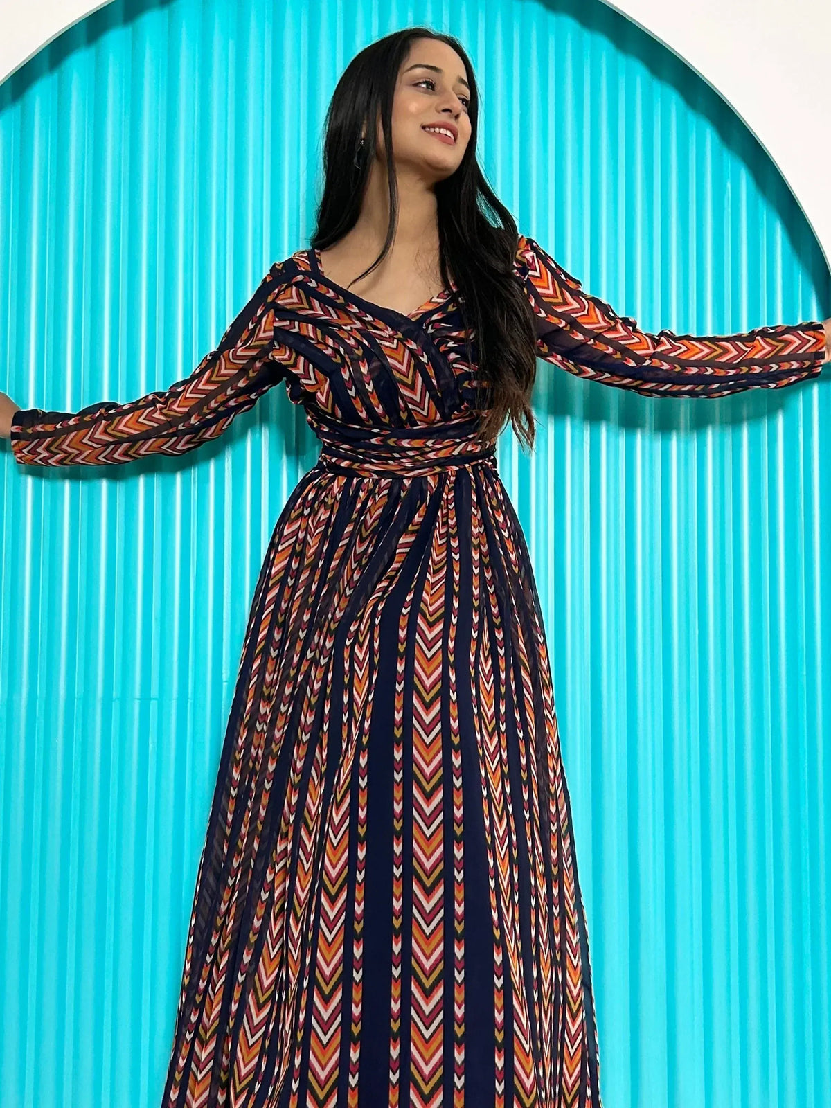 Woman in colorful ethnic print gown with long sleeves, standing against turquoise backdrop