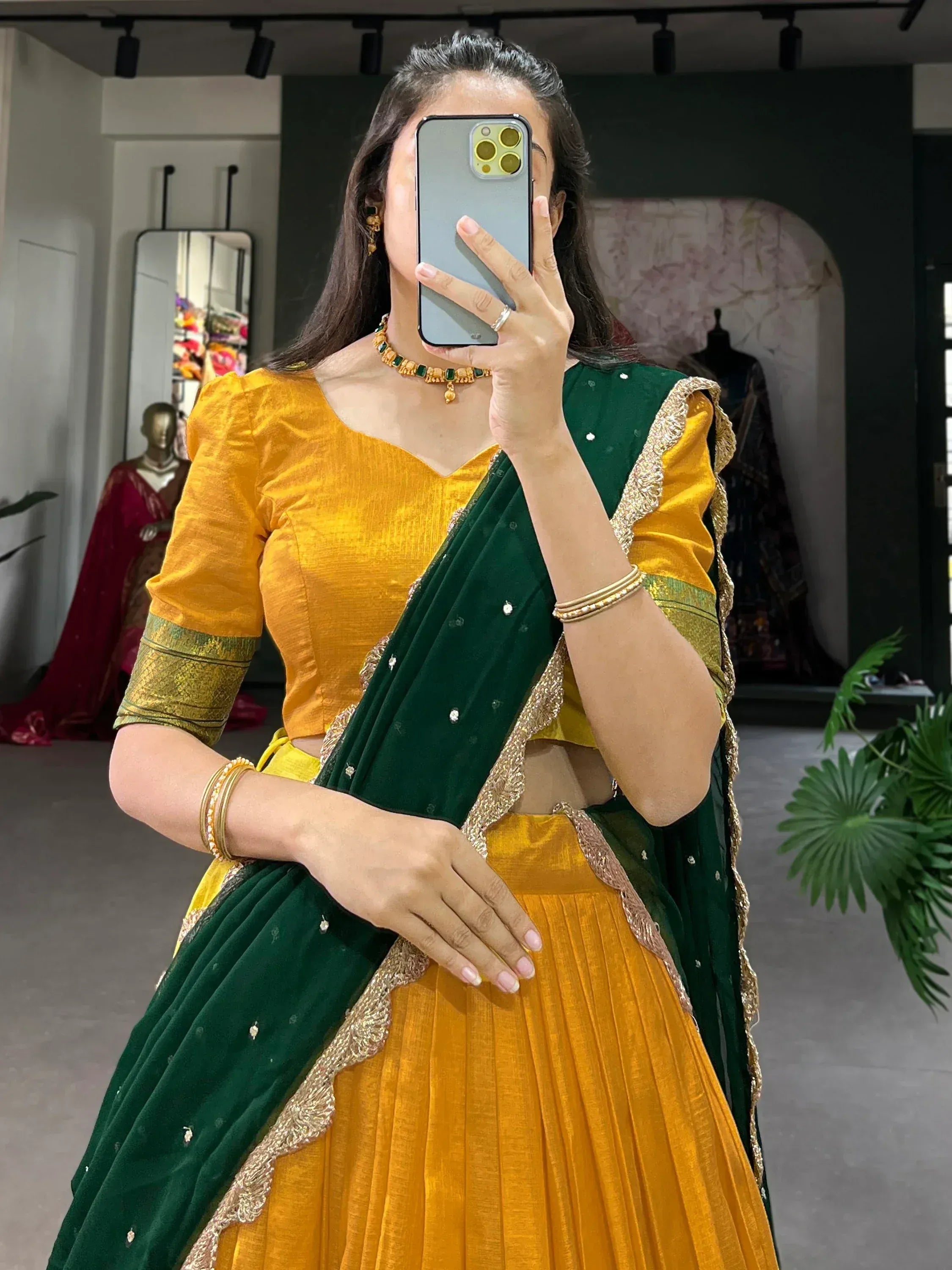 Woman in yellow and green designer lehenga choli with gold embroidery, taking mirror selfie in boutique