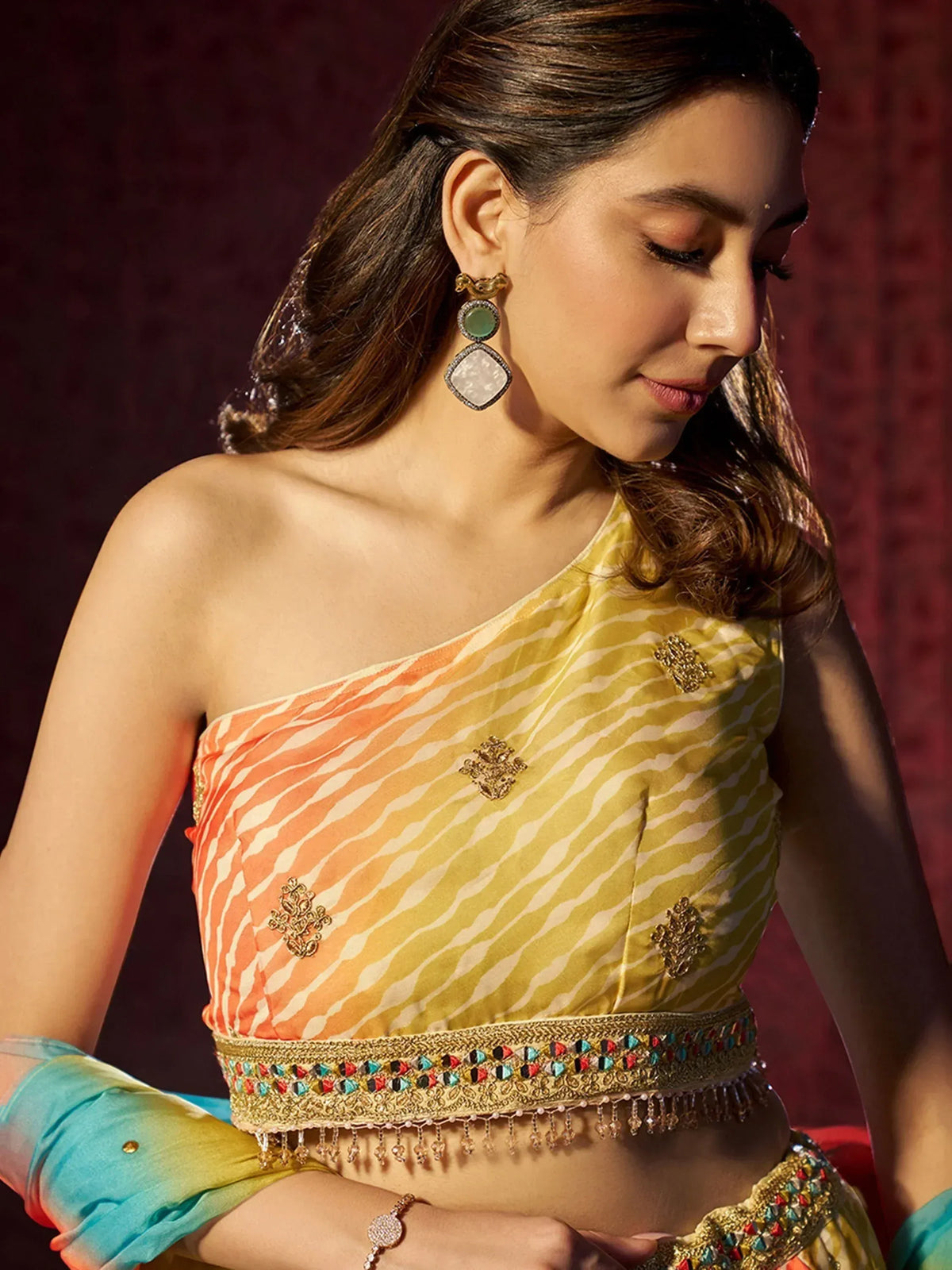 Woman in vibrant yellow and orange striped one-shoulder ethnic blouse with gold embroidery and colorful waistband, styled with statement earrings.
