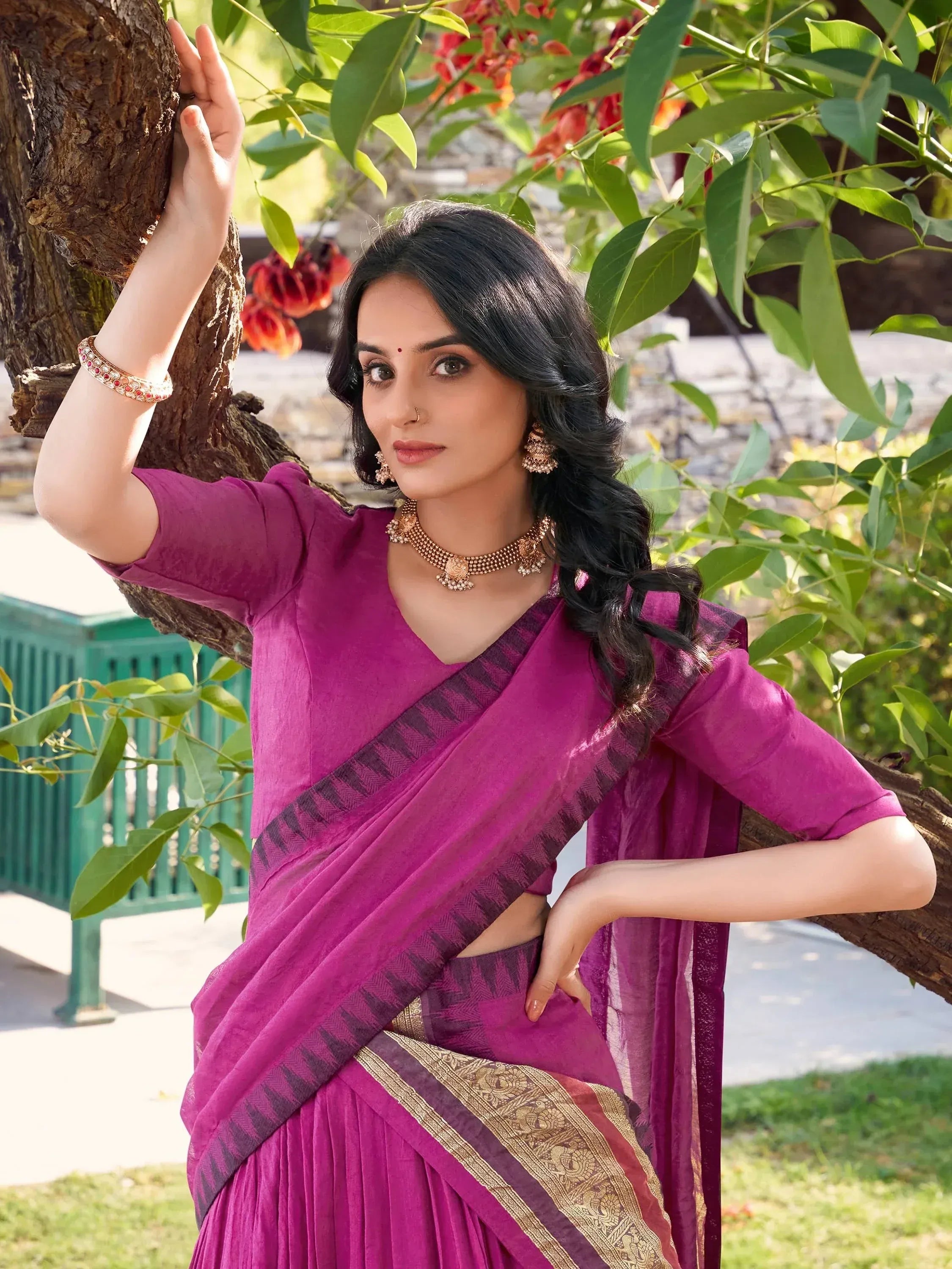 Woman in vibrant purple saree with gold border, styled with traditional jewelry, posing outdoors near tree